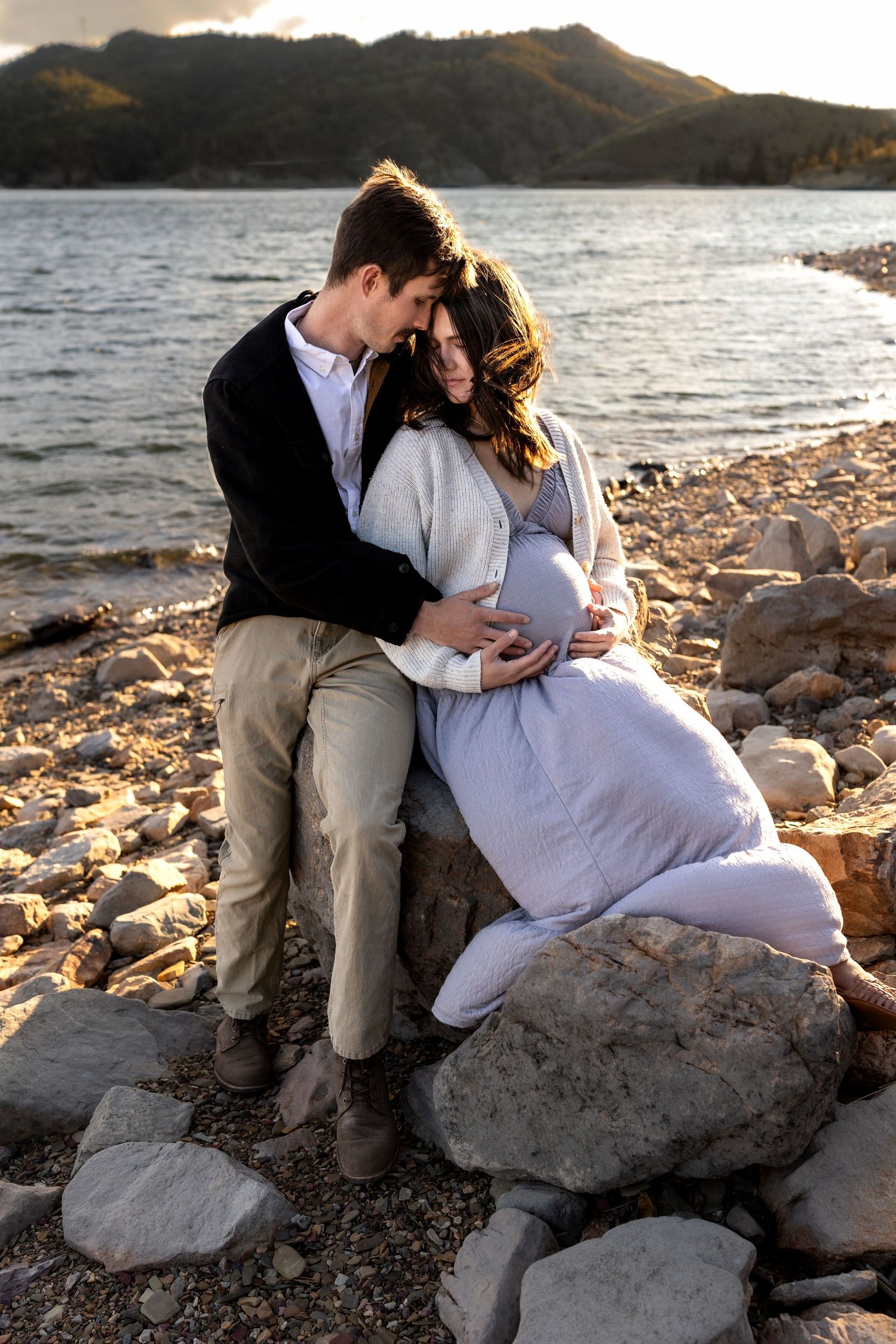 A mother and father rest their heads against each other as the sit on a rock in front of a lake and caress her pregnant belly