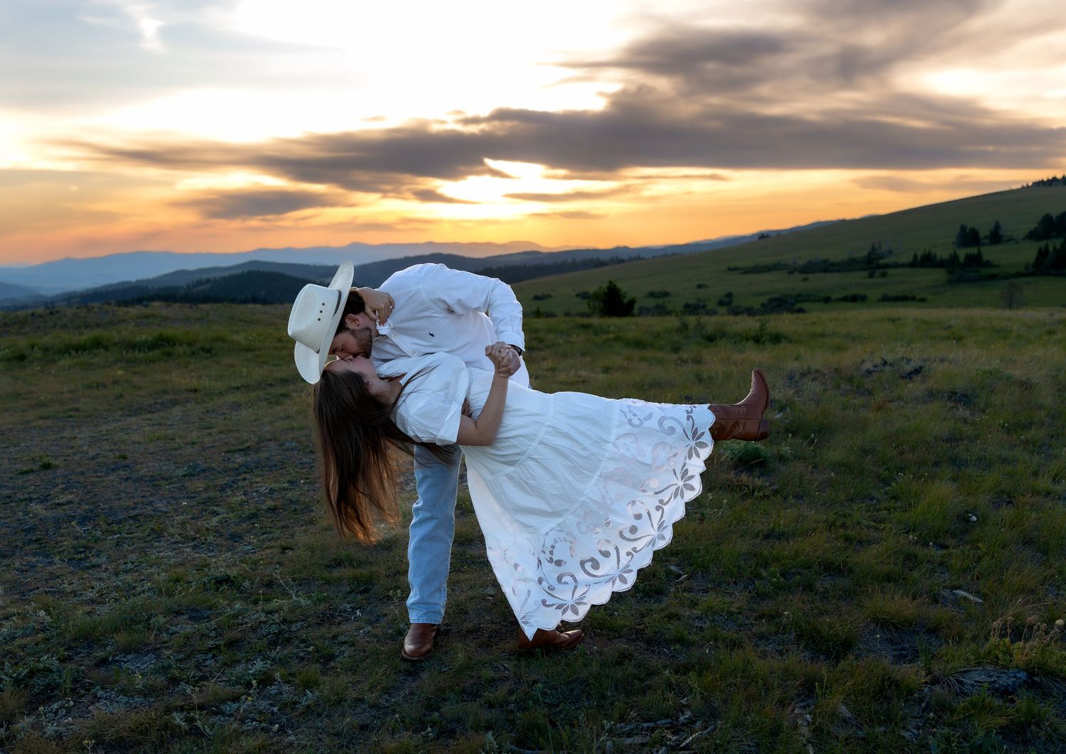 A man in a cowboy hat dips his fiancee and kisses her on a mountain top at sunset