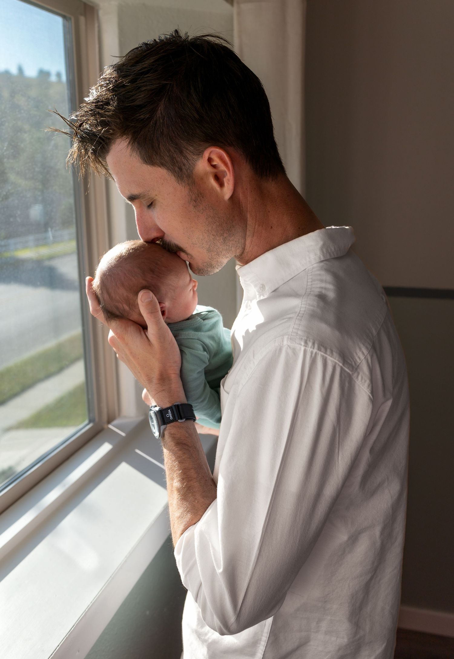 A new father kisses his sons head in his nursery in front of a window