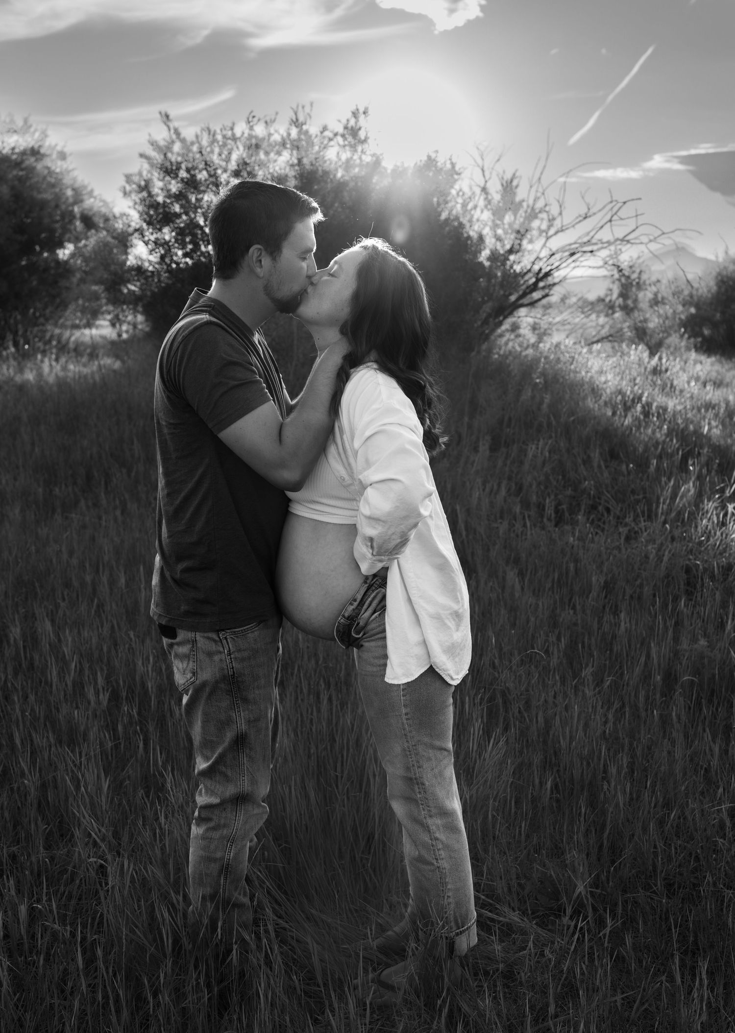 A black and white image of a pregnant woman and her husband sharing a kiss in a field surrounded by trees