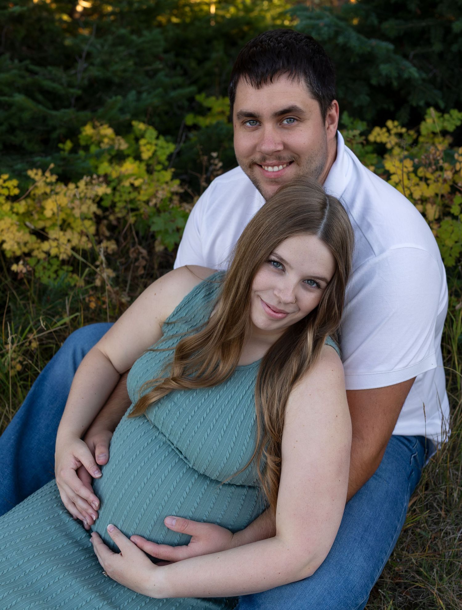 A man and his pregnant wife sit together in front of green and yellow bushes