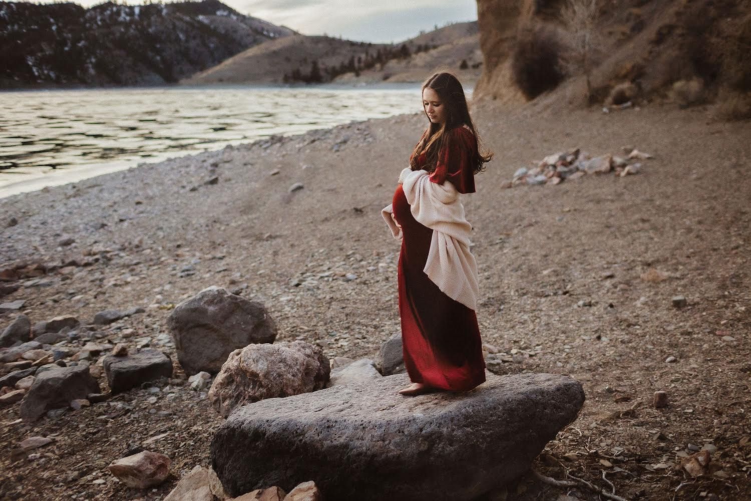 A woman caresses her pregnant belly as she stands on a rock by a lake