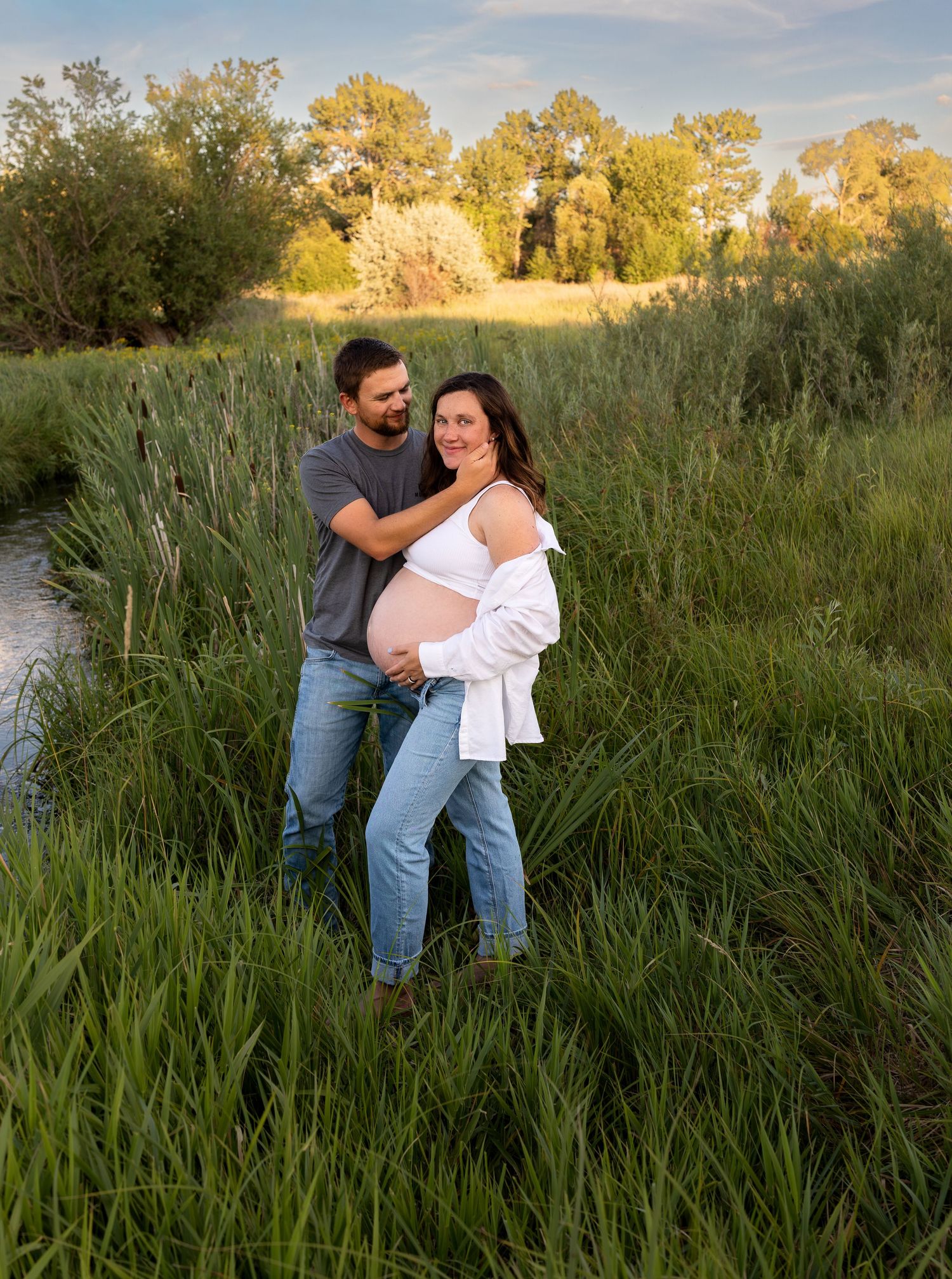 A Husband and Wife pose for a maternity photo in a lush green field.