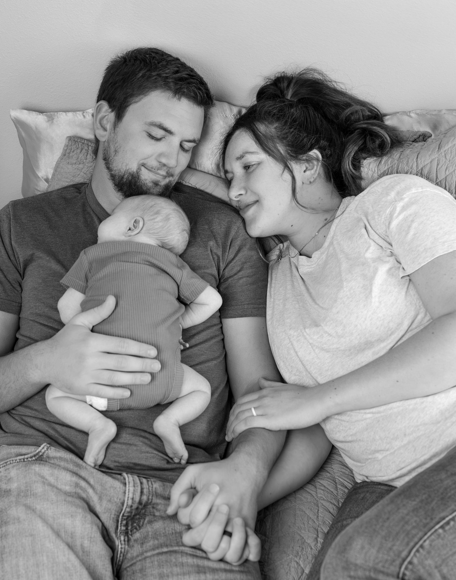 A Mom and Dad lay on their bed holding hands, with their newborn son sleeping on Dad's chest.