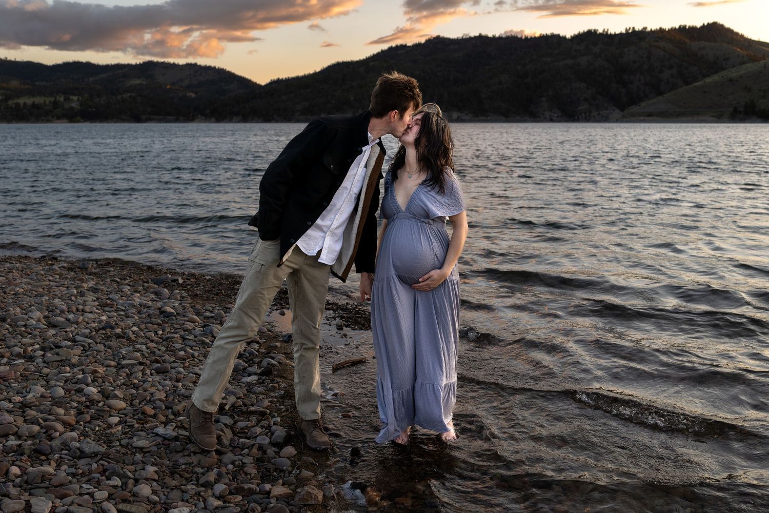 A pregnant woman walks along the shore of a lake as her husband leans over to kiss her