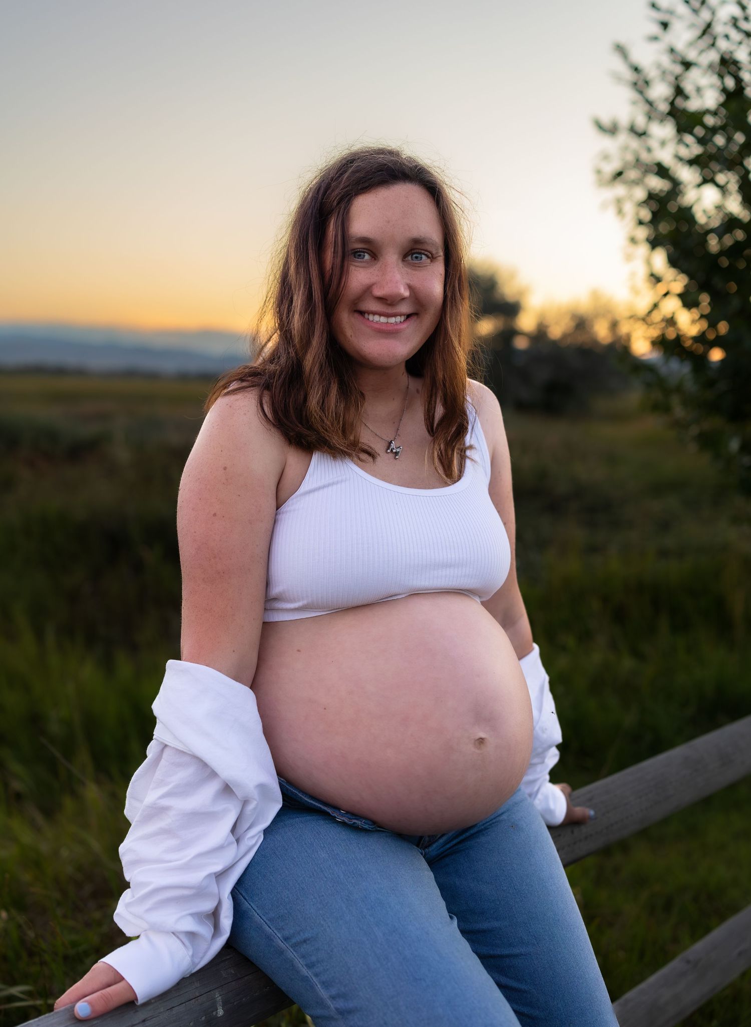 A pregnant woman sits on a wood fence at sunset