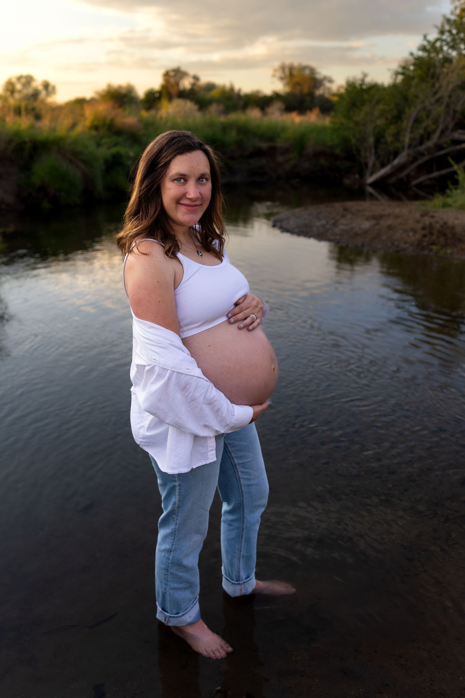 A pregnant woman holds her belly as she stands in a shallow creek at sunset