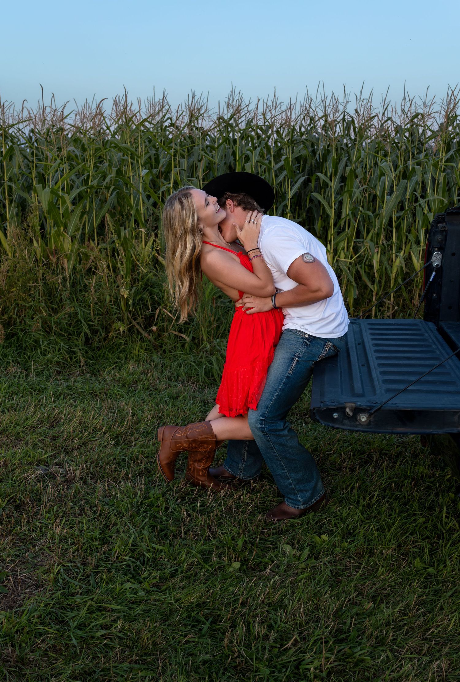 A man kisses a woman's neck on the tailgate of a truck in front of a corn field
