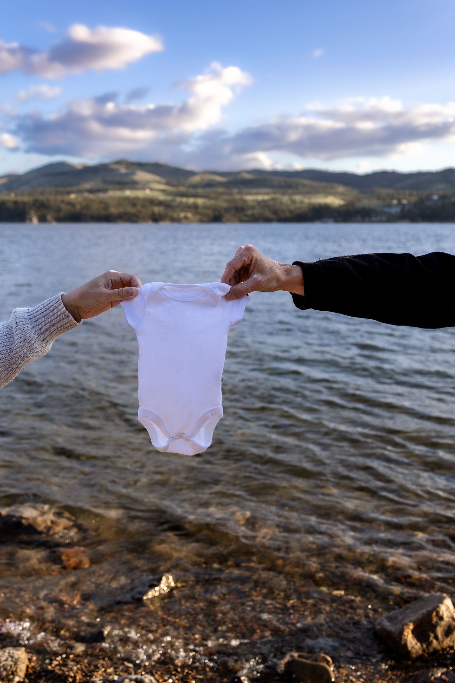 The hand of a mother and father hold up a white onesie in front of a lake and mountain