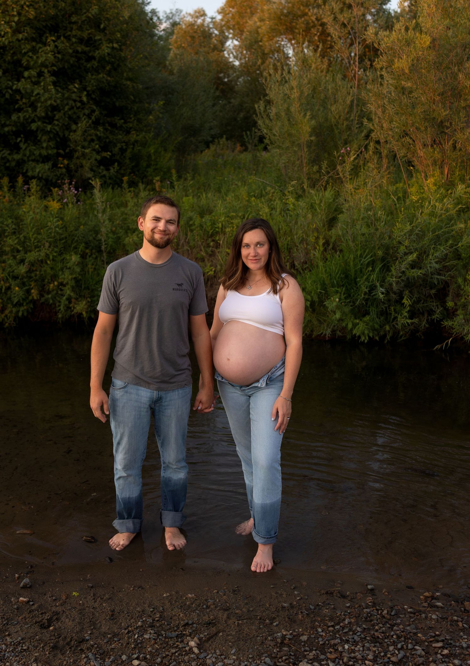 A man and his pregnant wife stand side by side in a shallow creek with green trees and bushes behind them