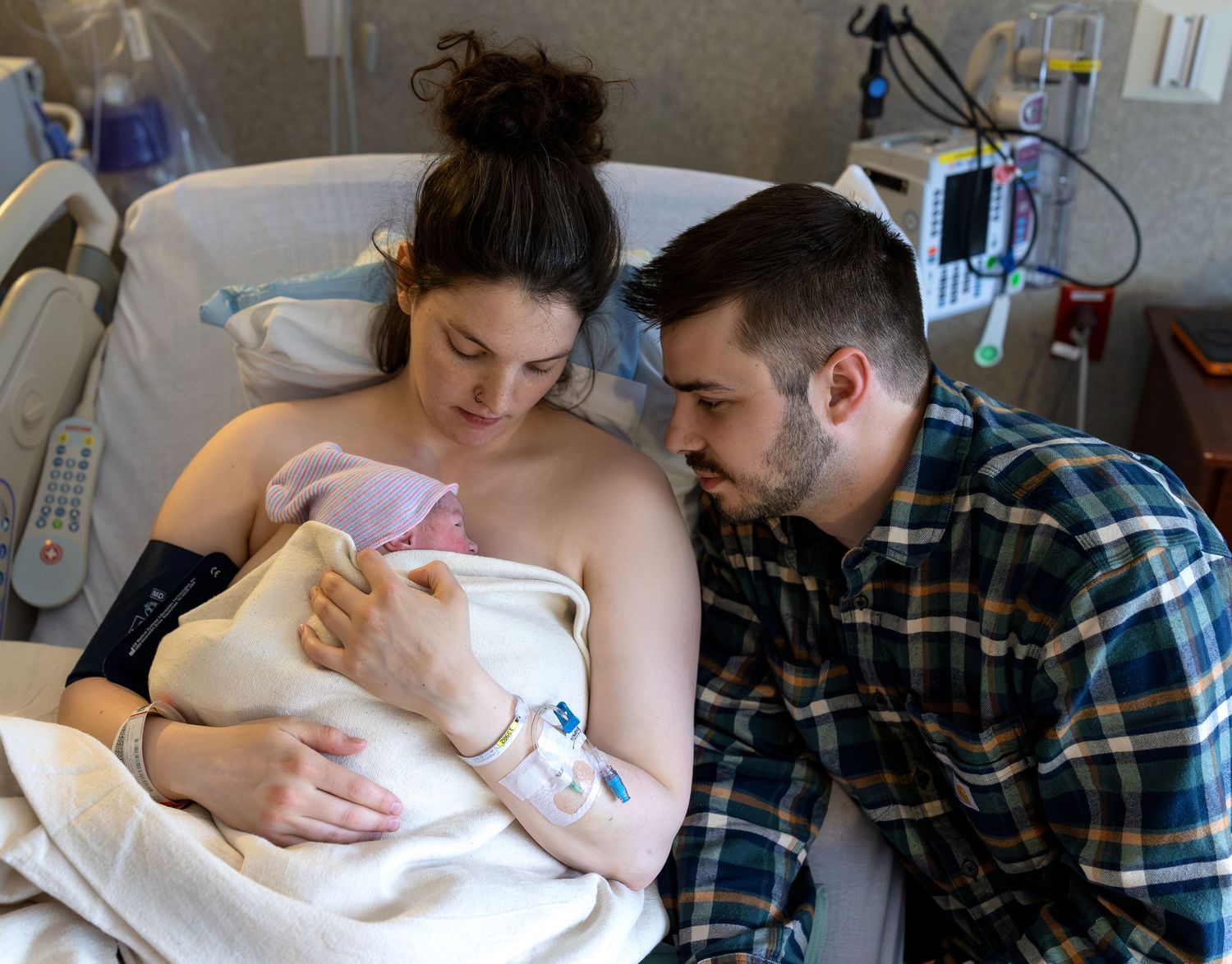 A mother sits in a hospital bed holding her newborn baby to her chest, the Father leans in from the side of the bed to look at his son.