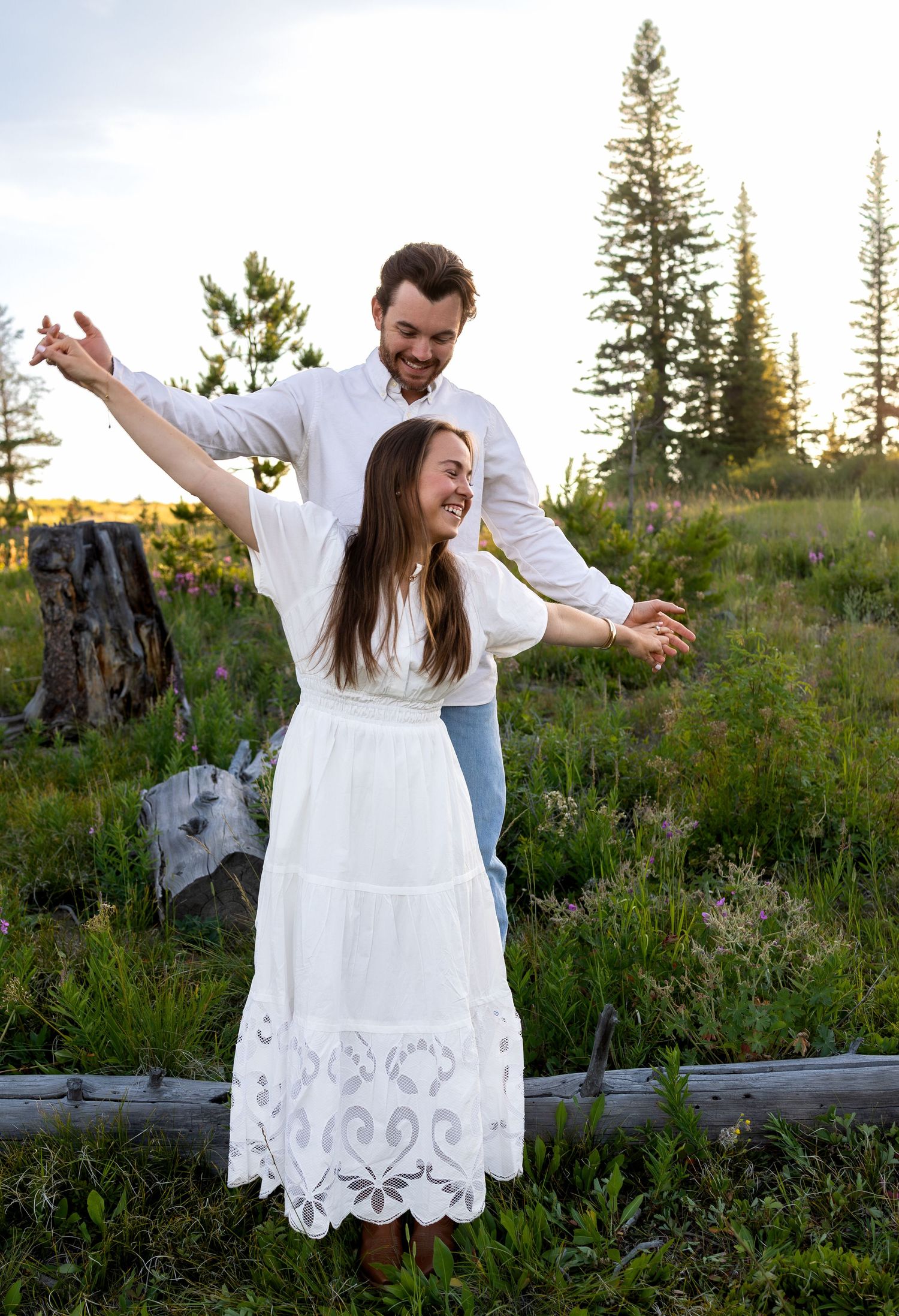A man and woman laugh as he stands on a log above his fiancee holding her hands