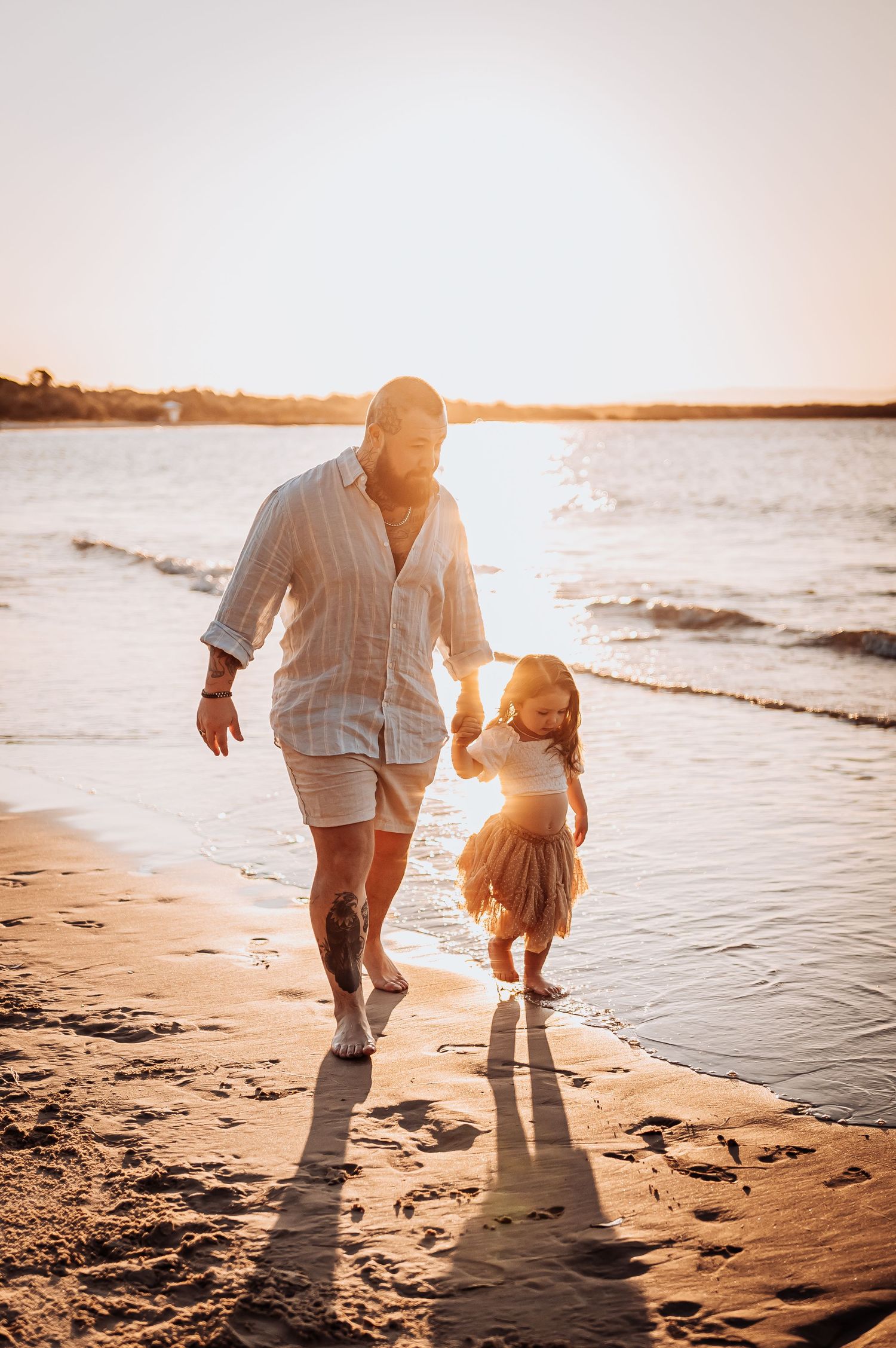 Parent and child walking together along beach shoreline at sunset casting long shadows.