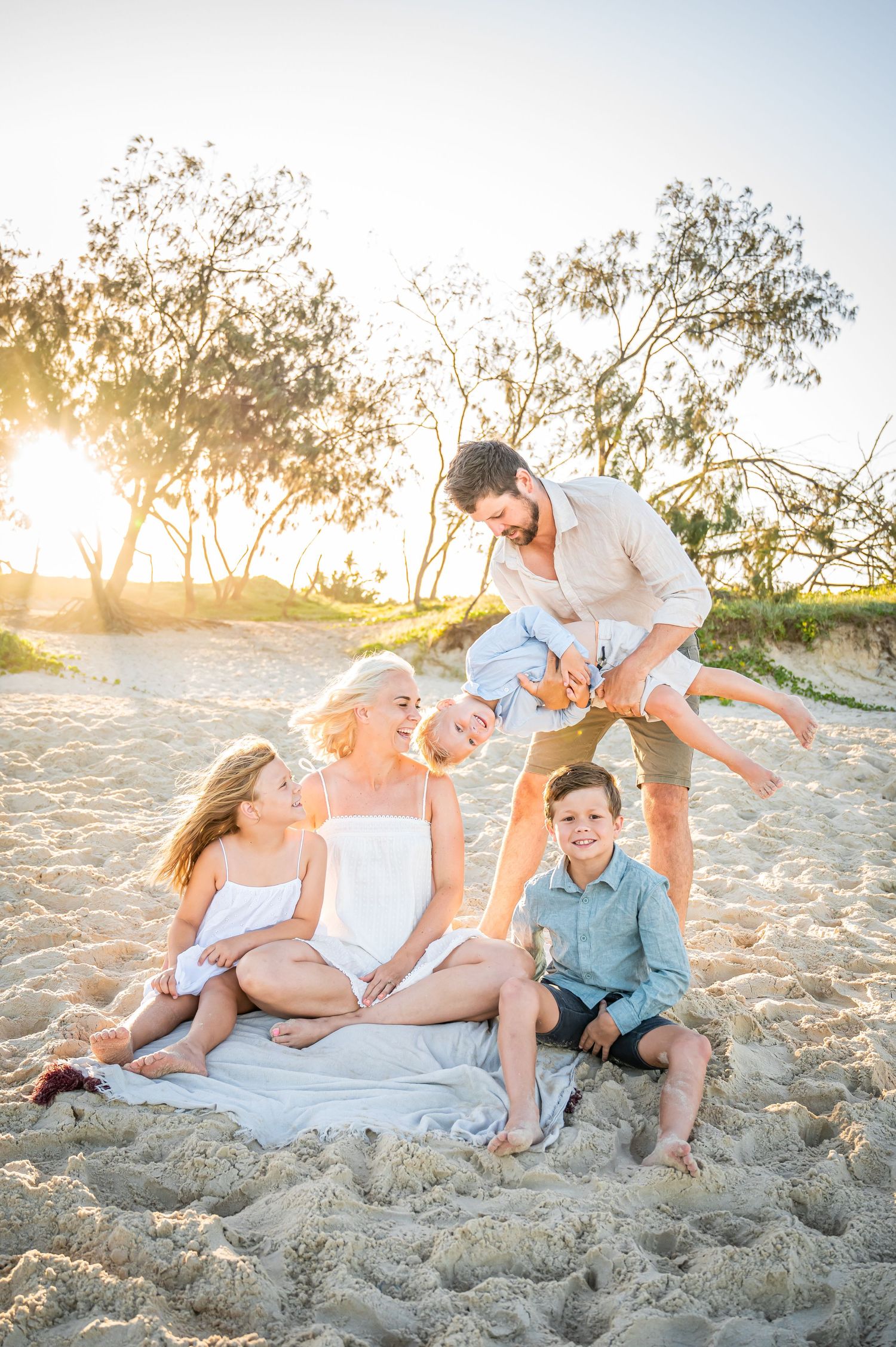 Group relaxes on blanket at beach during golden hour with trees and sand dunes in background.