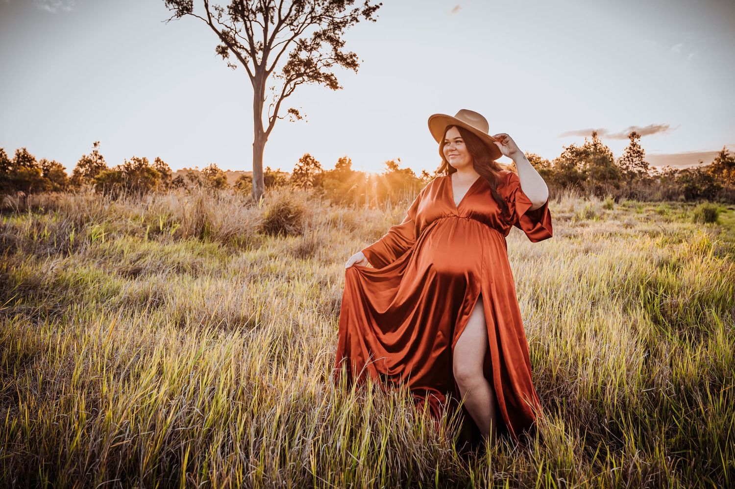 Maternity portrait in flowing rust-colored dress amid golden grass at sunset.