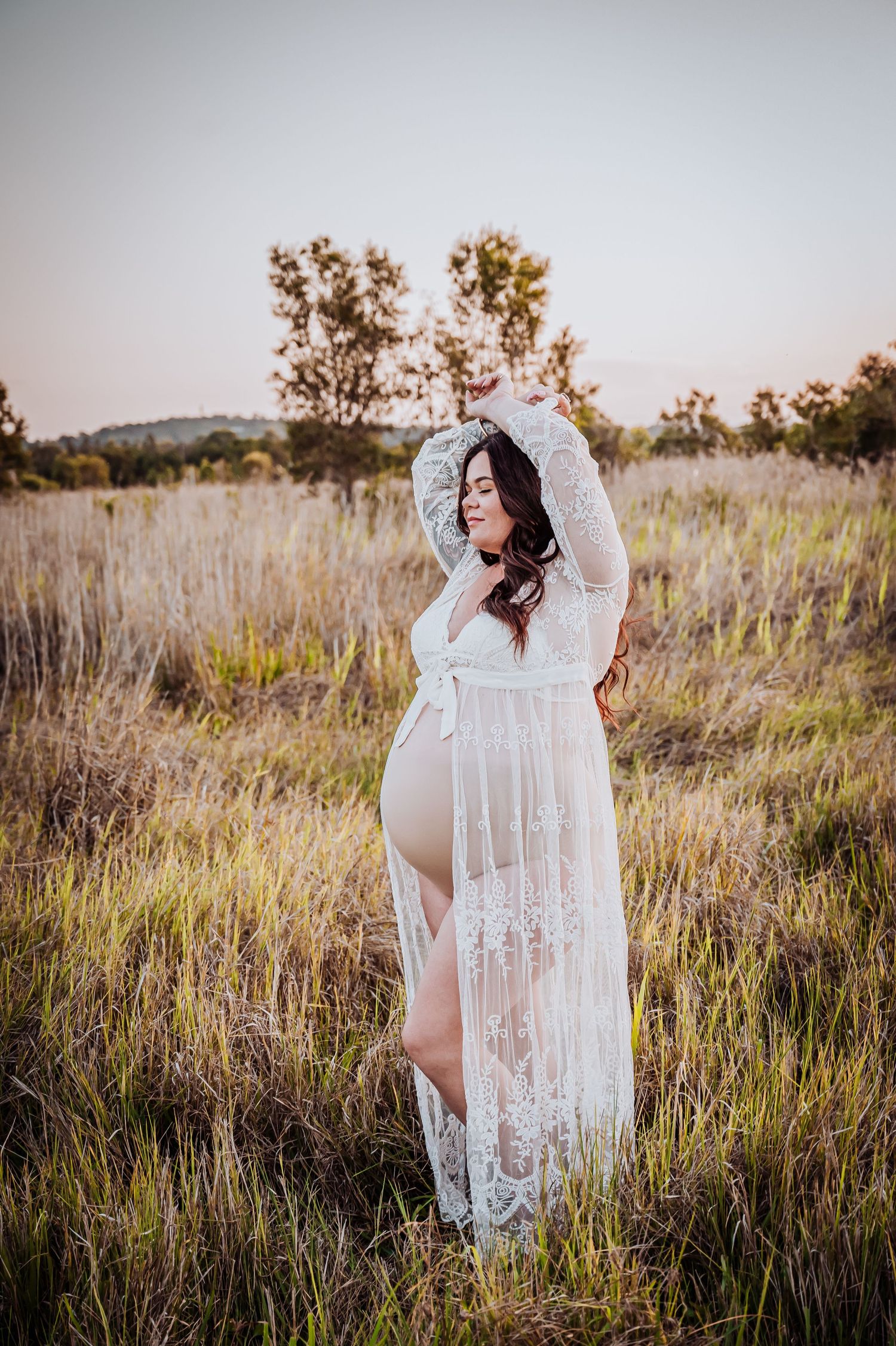 Ethereal maternity portrait in a white lace dress standing in tall grass at golden hour.