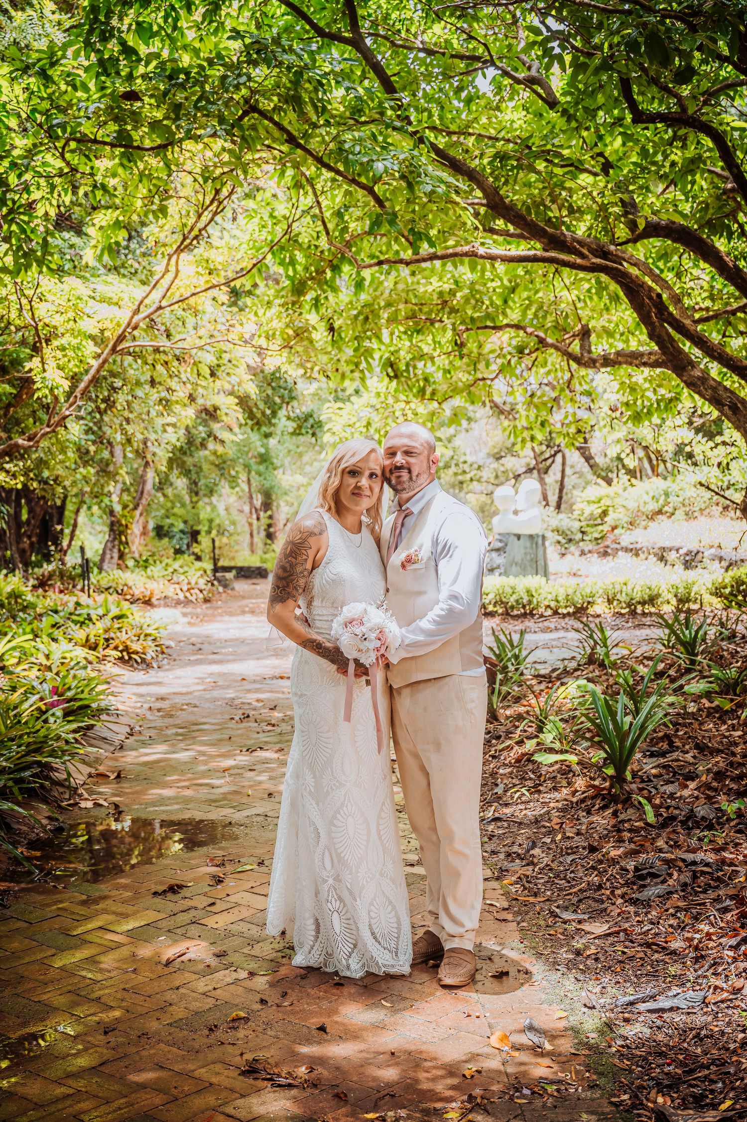 Wedding couple posing on garden path under lush green tree canopy.