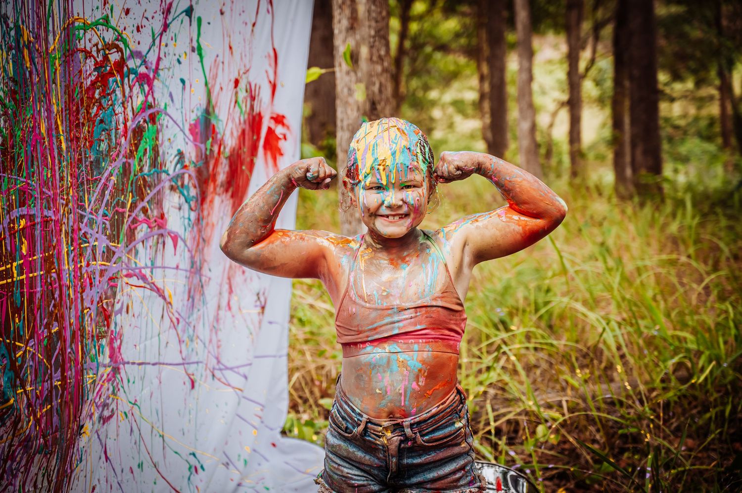 Child covered in colorful paint splashes poses joyfully against paint-splattered backdrop.