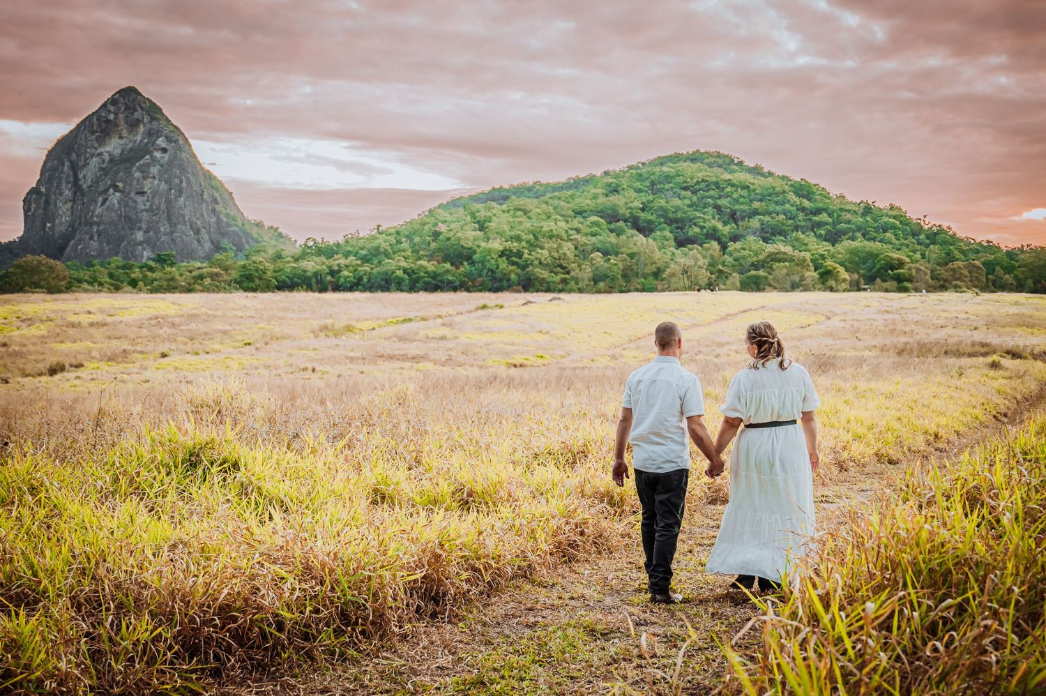 A couple walks through a golden field with a dramatic mountain peak looming in the background.