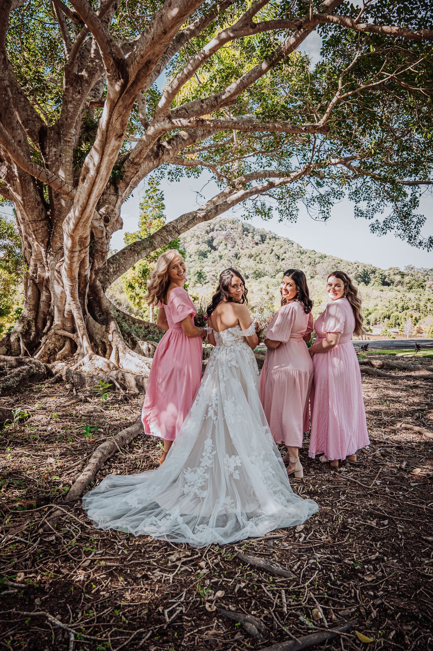 Wedding party gathered under a large tree with bridesmaids in pink dresses surrounding a bride in a lace gown.