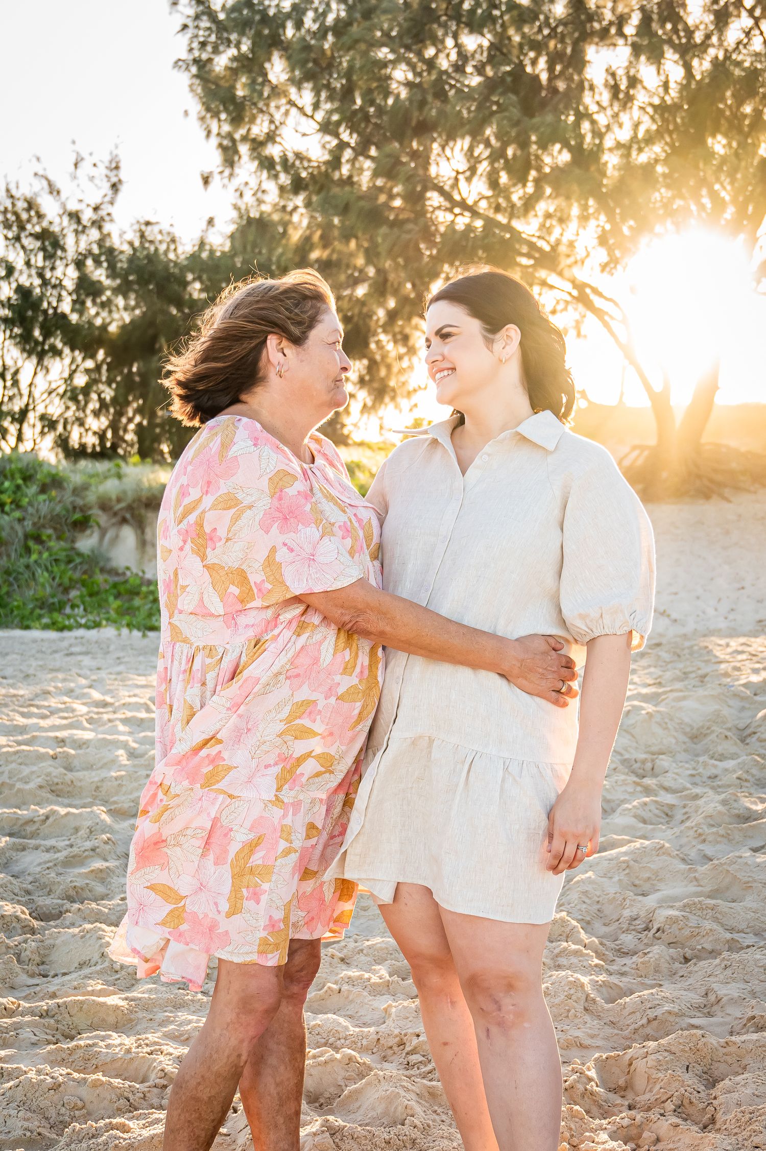 Two people share an intimate moment on beach at sunset with golden light filtering through trees.