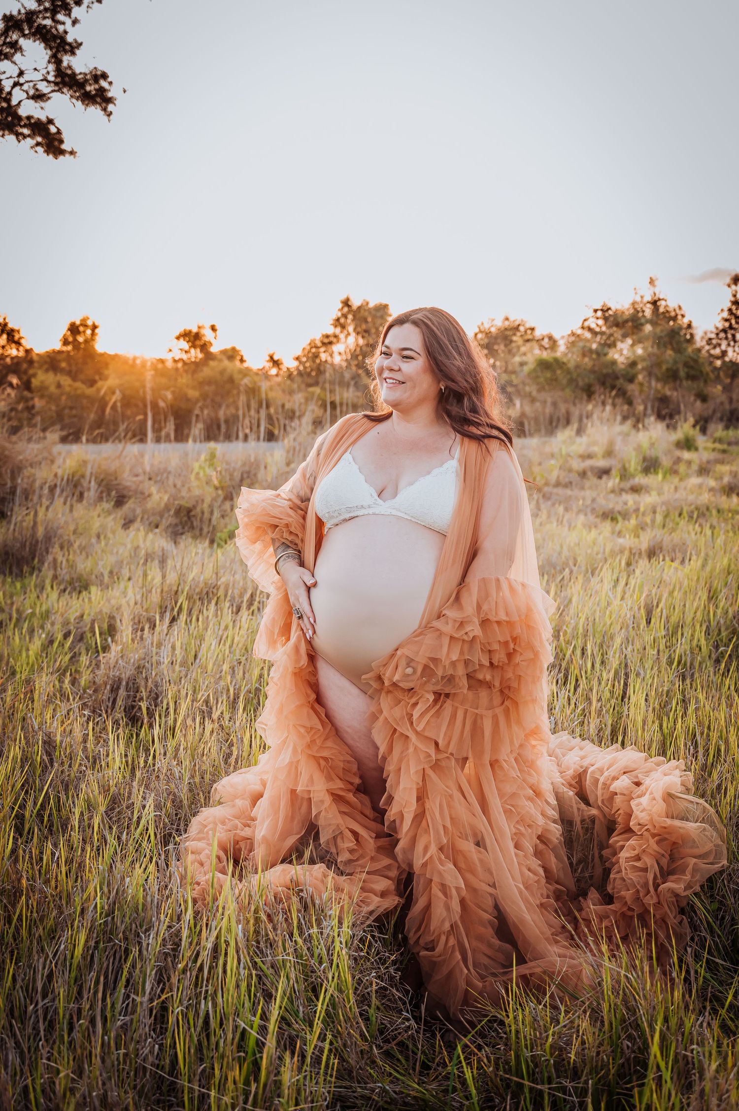 Dreamy maternity portrait in peach tulle gown amid tall grass at golden hour.