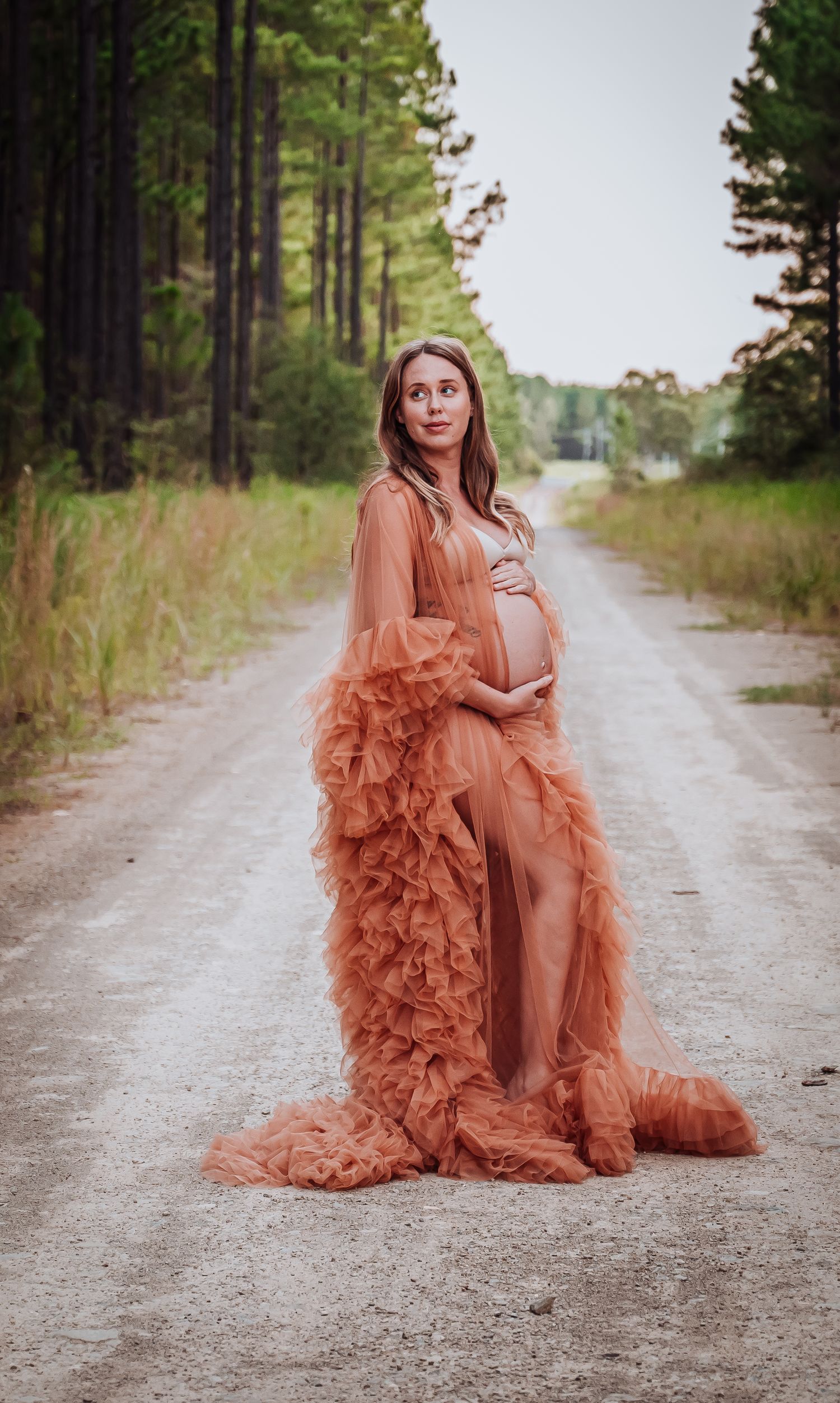Dramatic maternity portrait on a dirt road with flowing coral dress and pine forest backdrop.