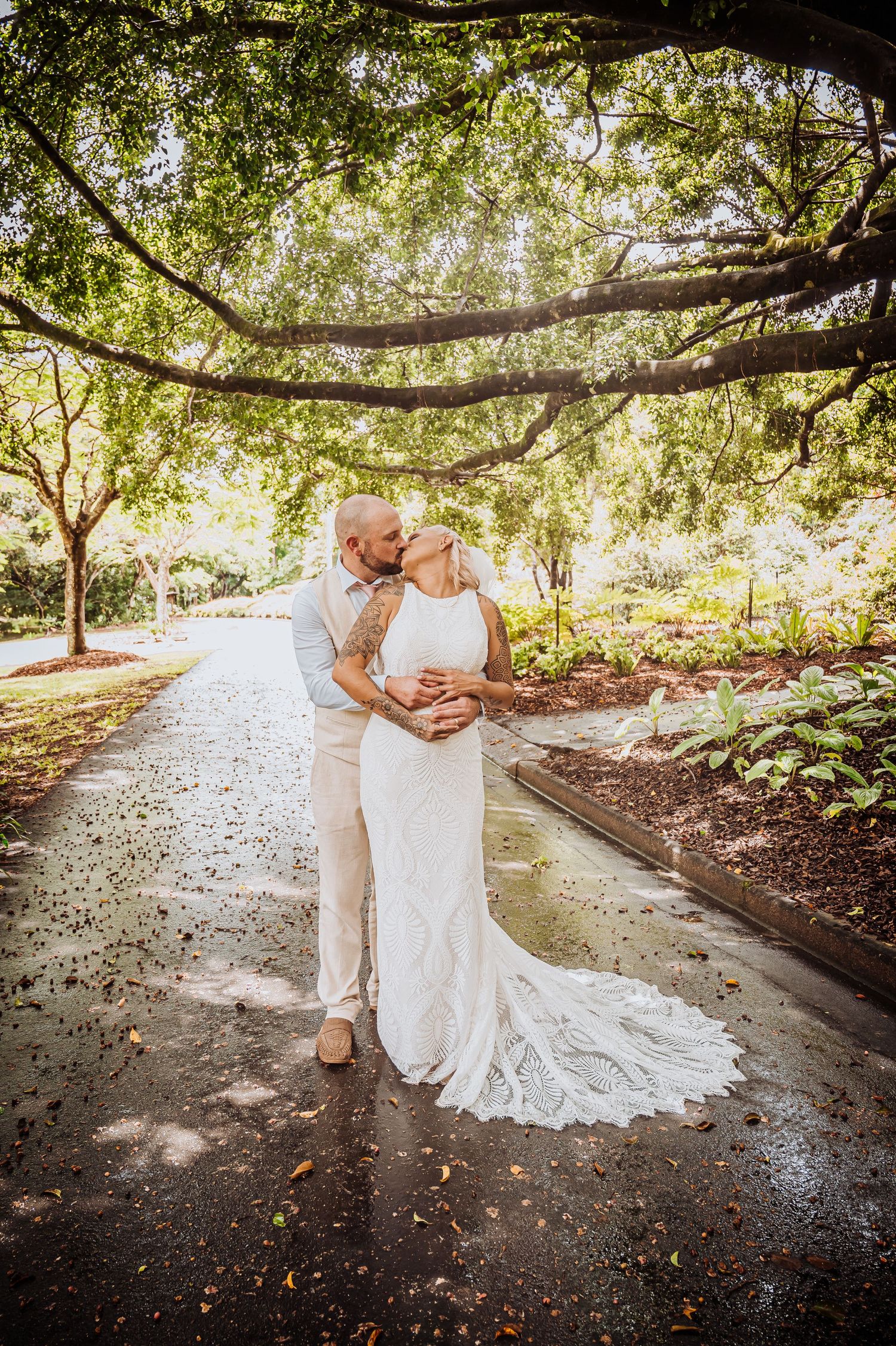 Romantic embrace under tree canopy on garden path with flowing wedding dress train.