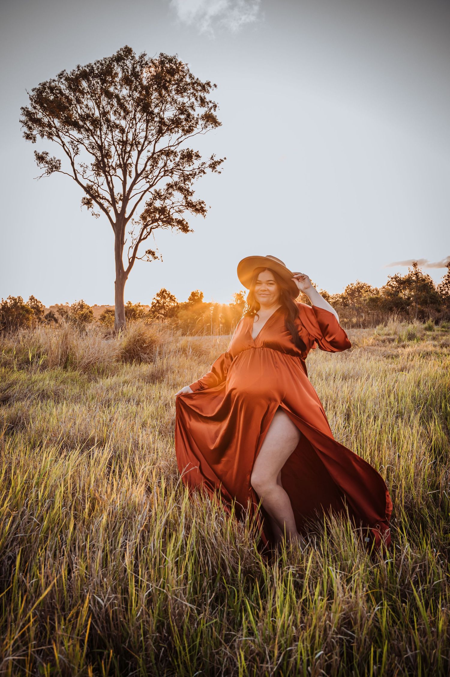 Woman in flowing rust-colored dress twirls in tall grass at sunset with lone tree in background.