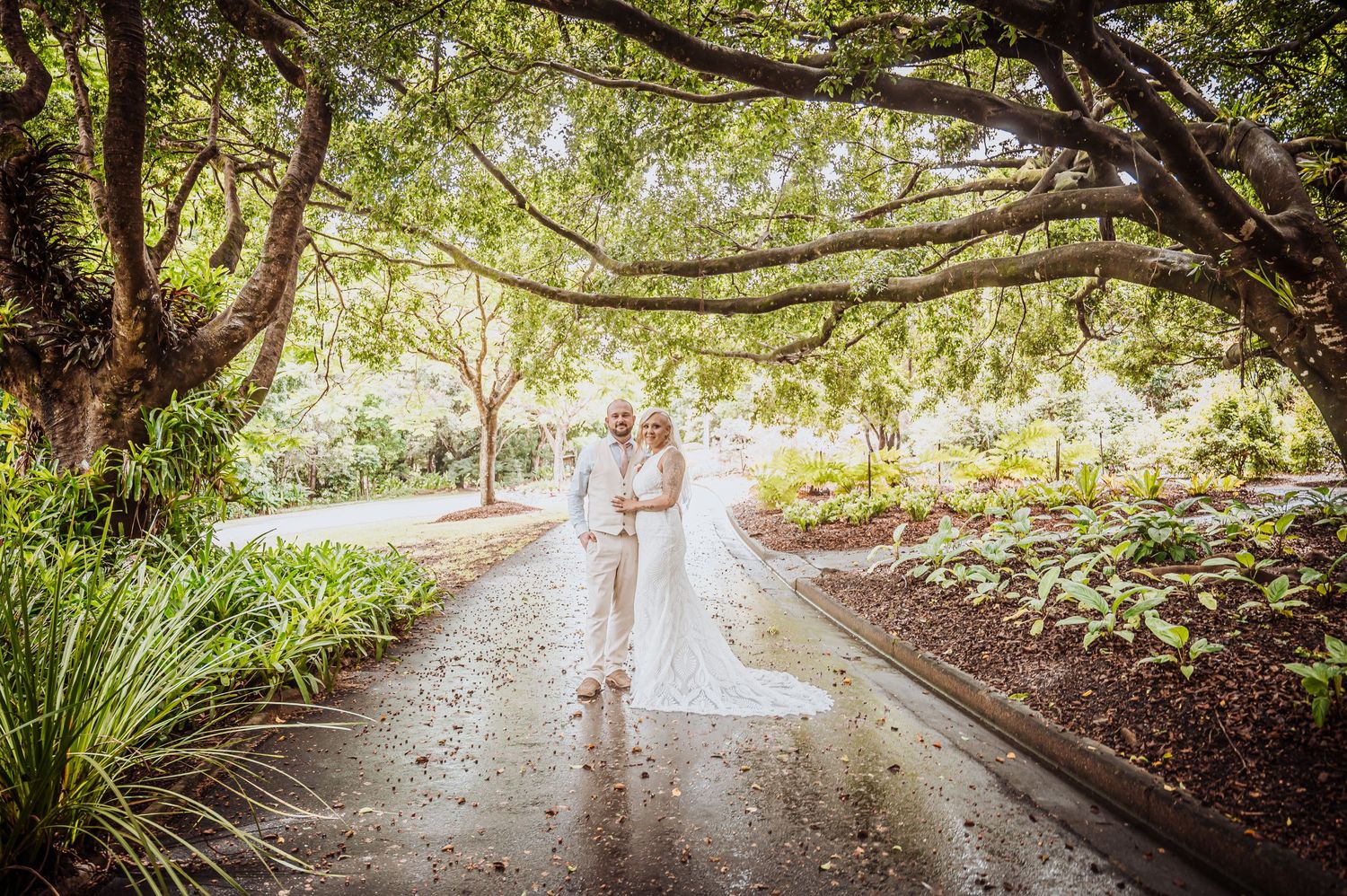 Ethereal wedding couple pose together on a garden path beneath overhanging tree branches.