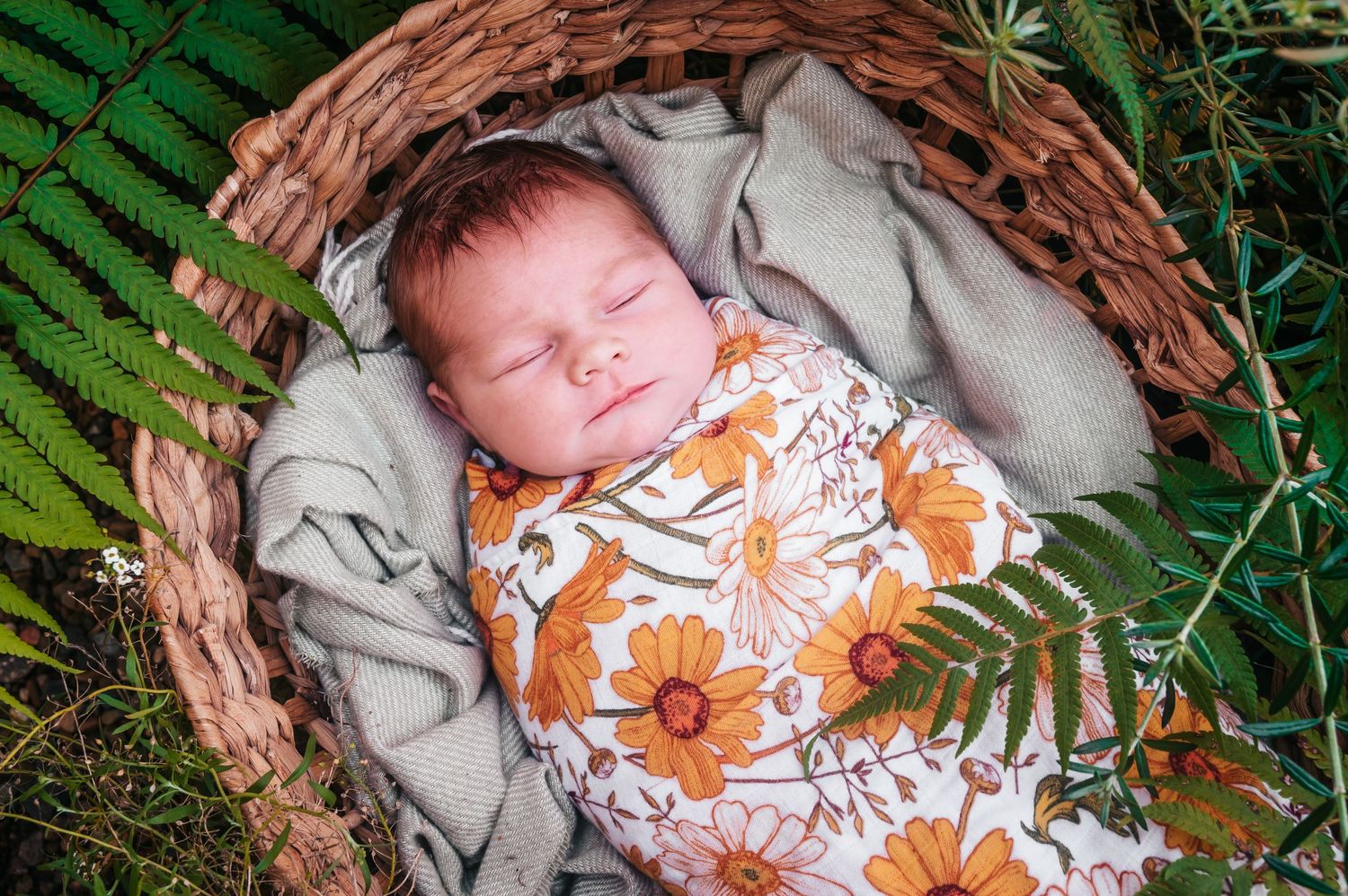 Peaceful newborn baby sleeping in wicker basket with floral swaddle and fern accents.