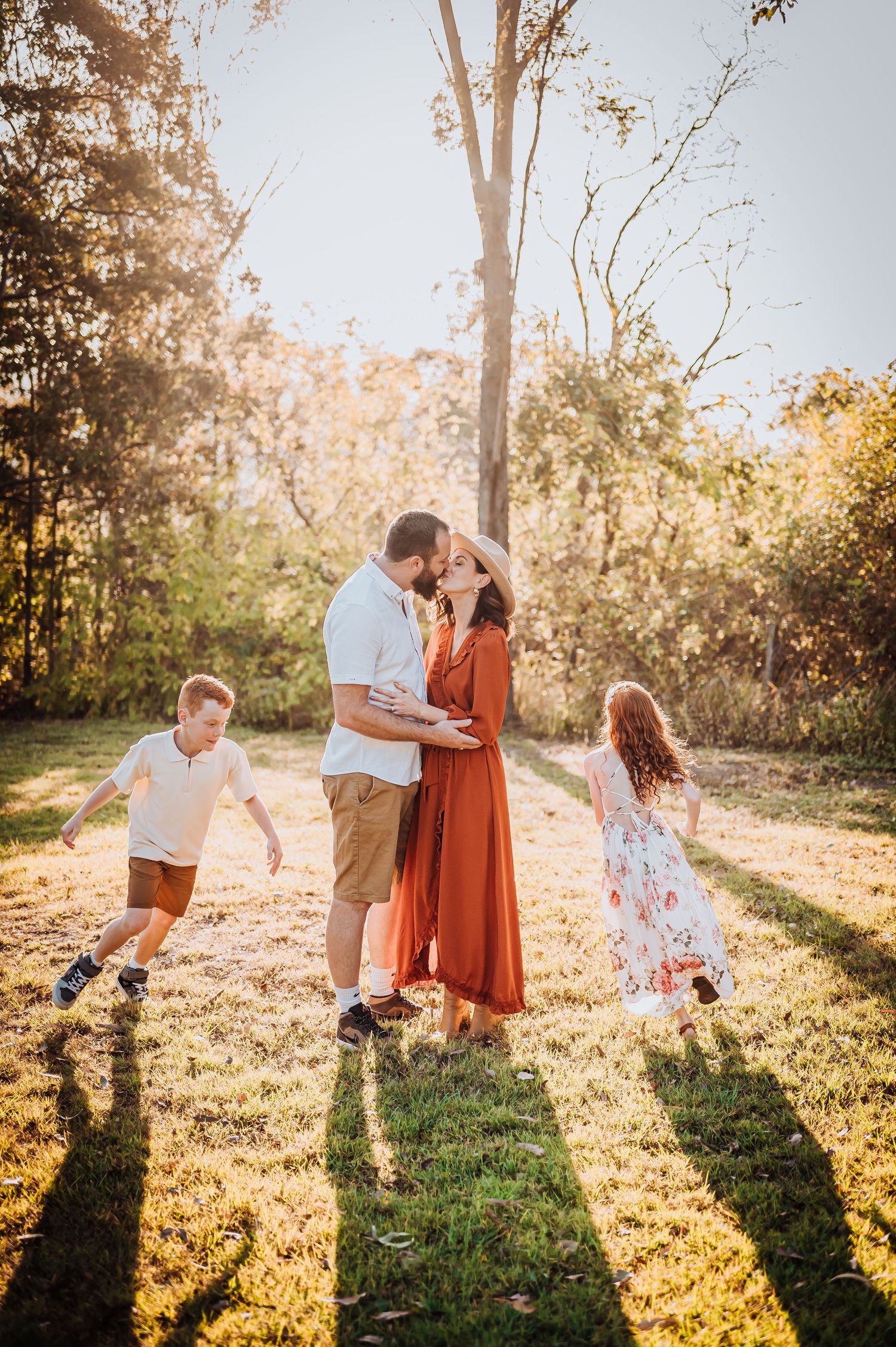 A family enjoys a golden sunset in a grassy field as children play and run around their embracing parents.