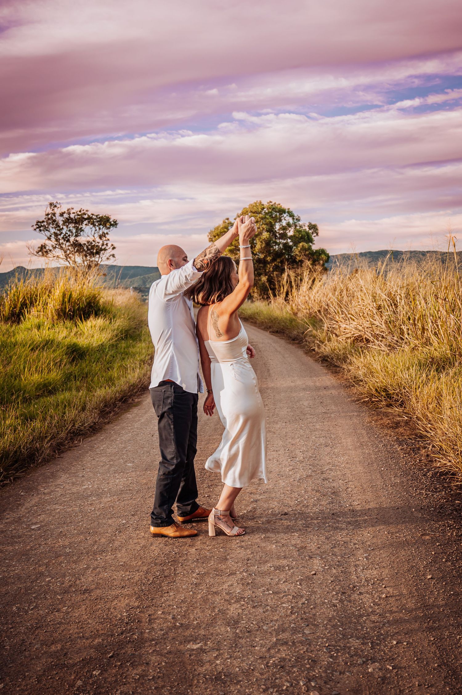 A couple dances together on a dirt road surrounded by golden grass at sunset.