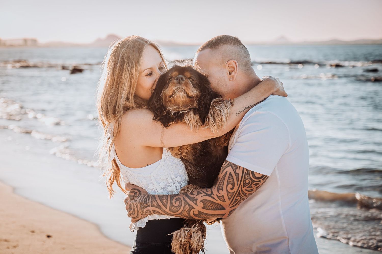 A couple shares an intimate embrace on a beach with their dog at sunset.