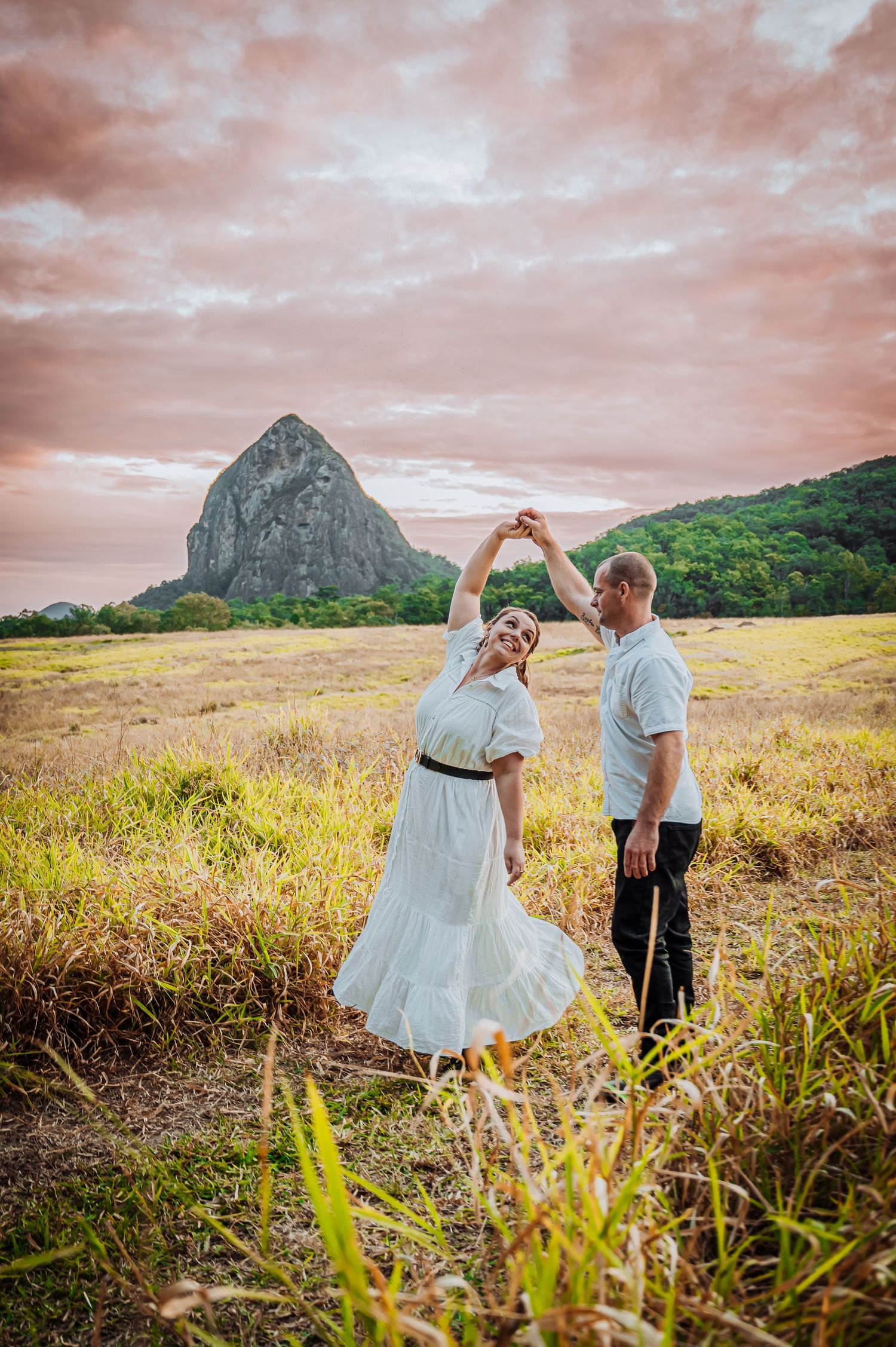 A couple dances in a field with a prominent mountain peak and pink sunset sky in the background.