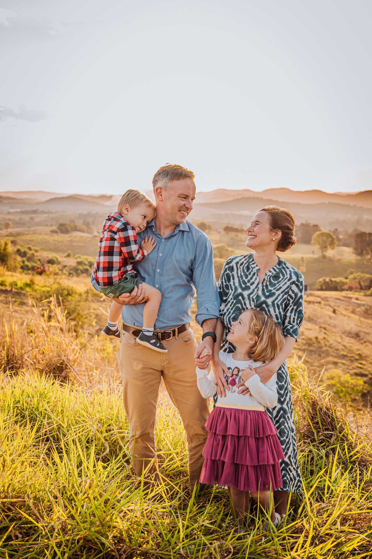 Family shares loving moment together in golden grassland with rolling hills in background.
