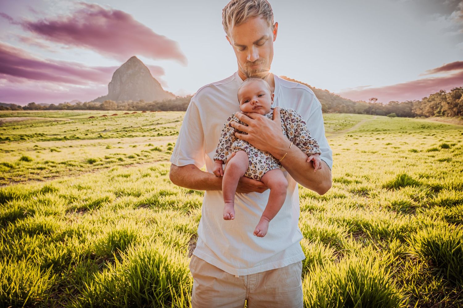 Adult cradling infant in white outfit against backdrop of mountains and sunset.