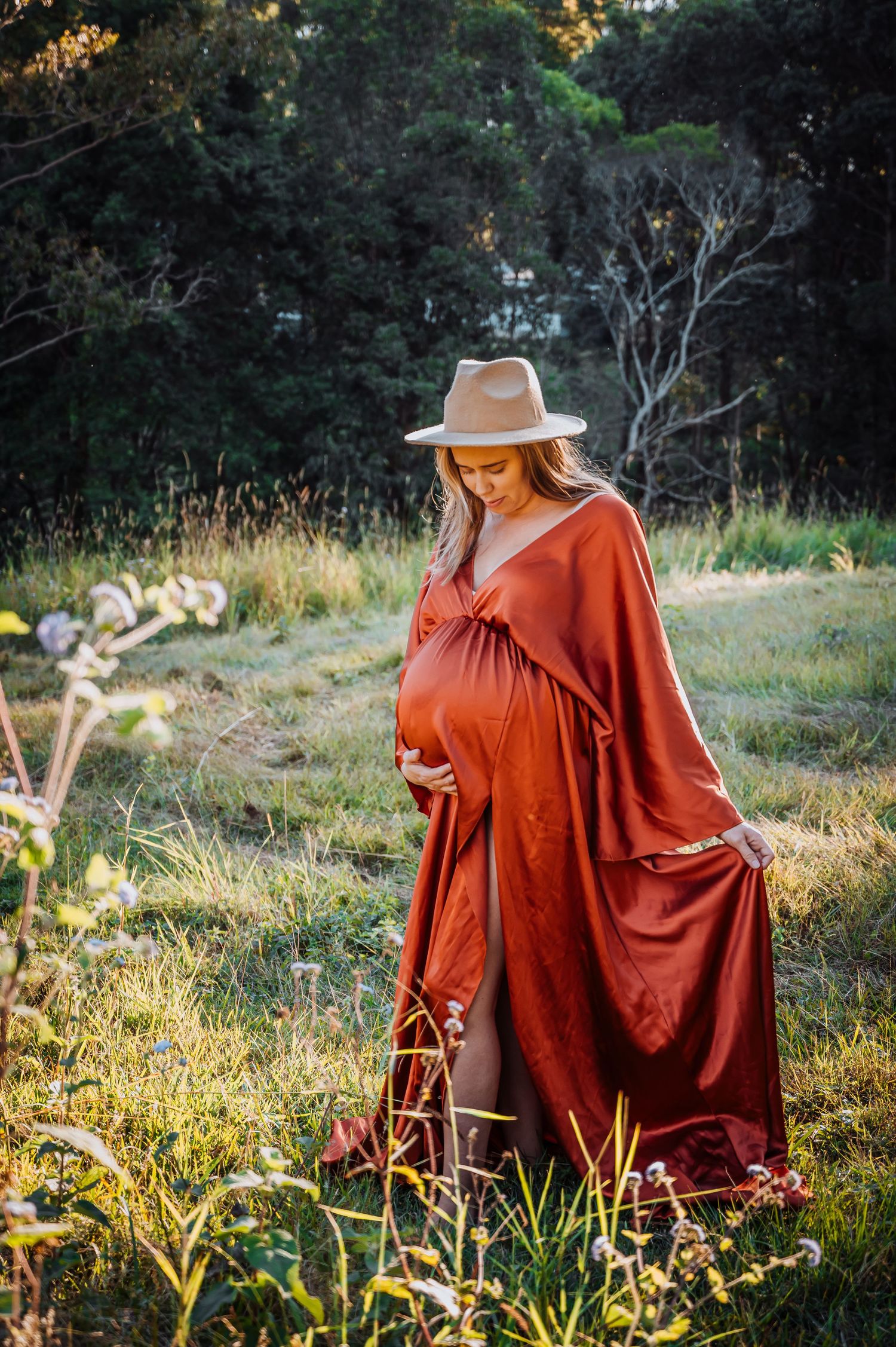 Maternity portrait in flowing rust-colored dress and hat among wildflowers at sunset.