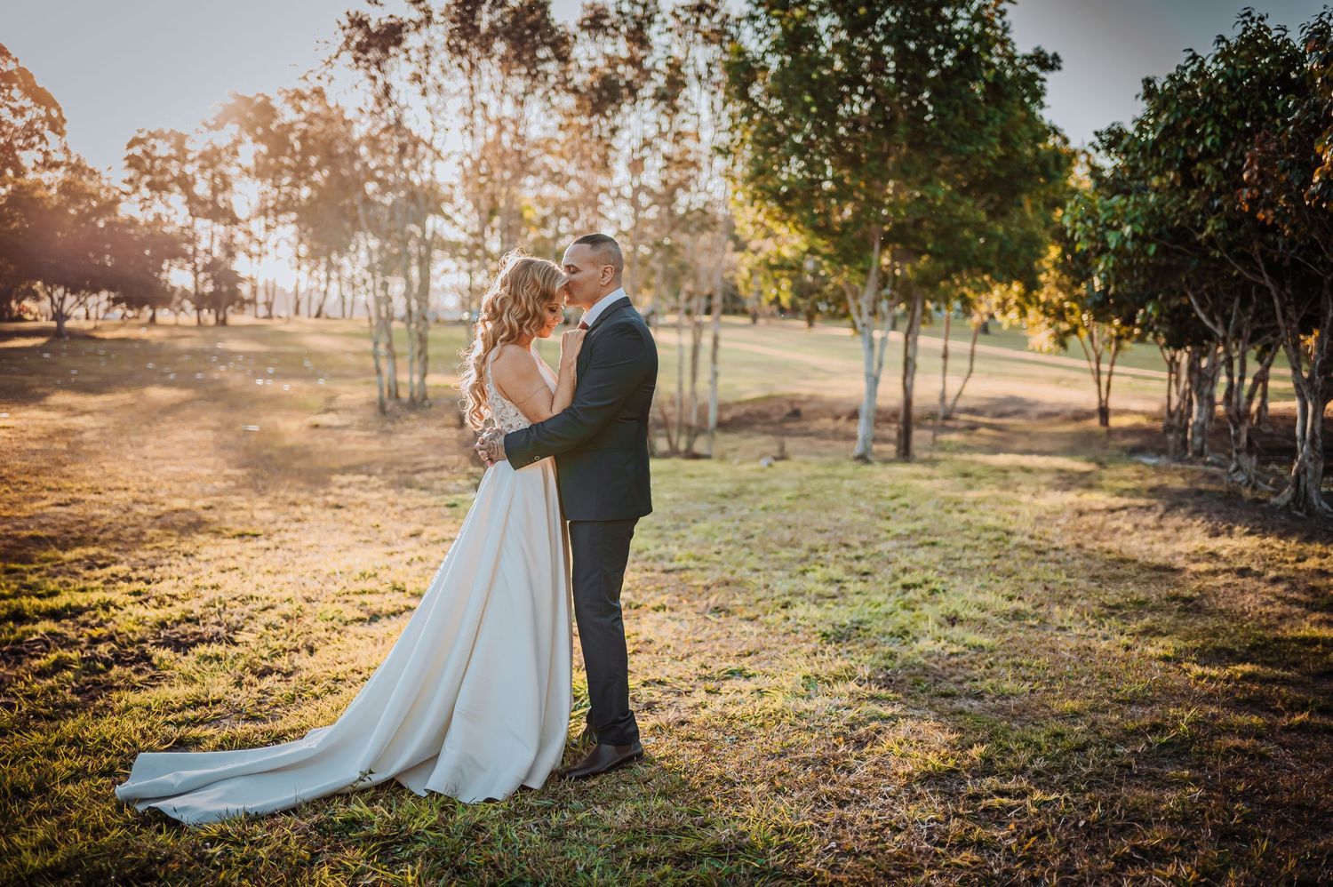 Wedding couple share intimate moment in sunlit field with trees in background.