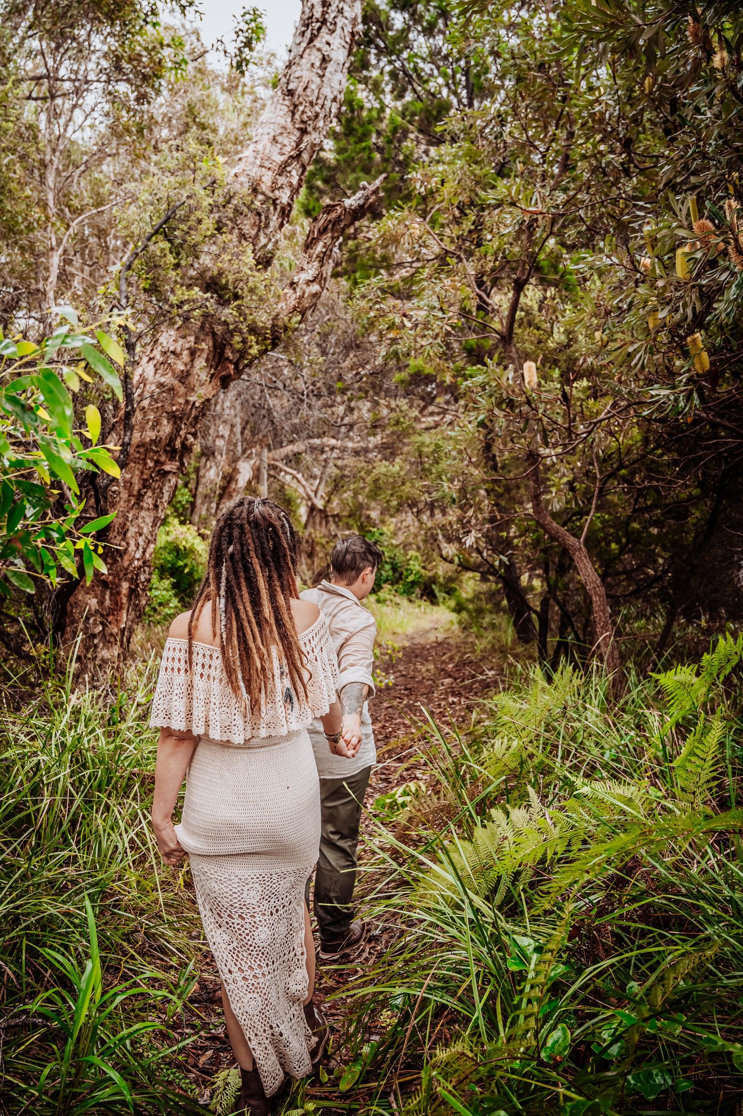 A couple walks hand in hand along a nature trail surrounded by green foliage.
