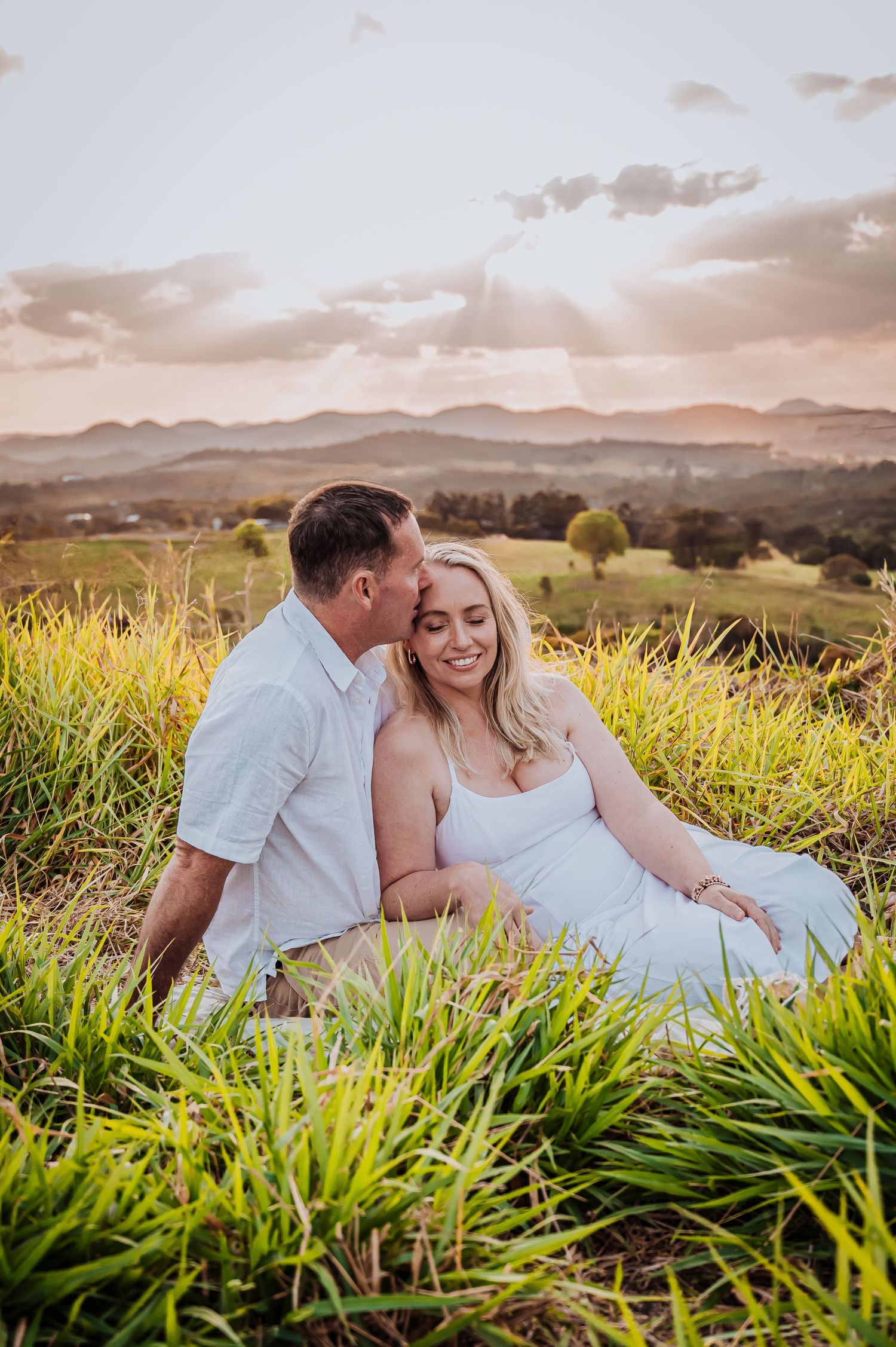 A couple sits together in tall grass during golden hour with mountains visible in the distance.