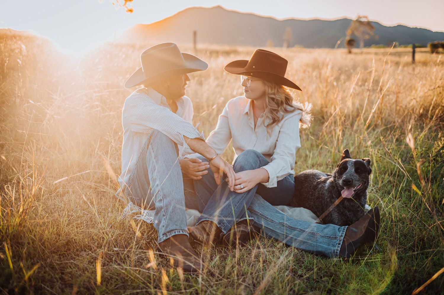 A couple in western wear sits in a sunlit field with their dog during golden hour.