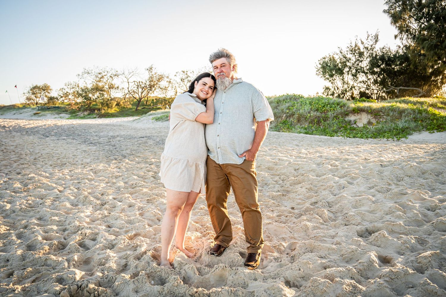 Couple poses for portrait on sandy beach during golden hour with coastal vegetation behind.