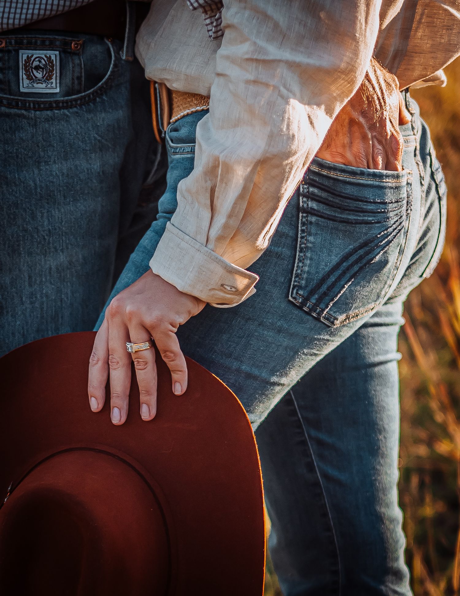 Close-up detail of denim jeans and a hand holding a brown cowboy hat in warm lighting.