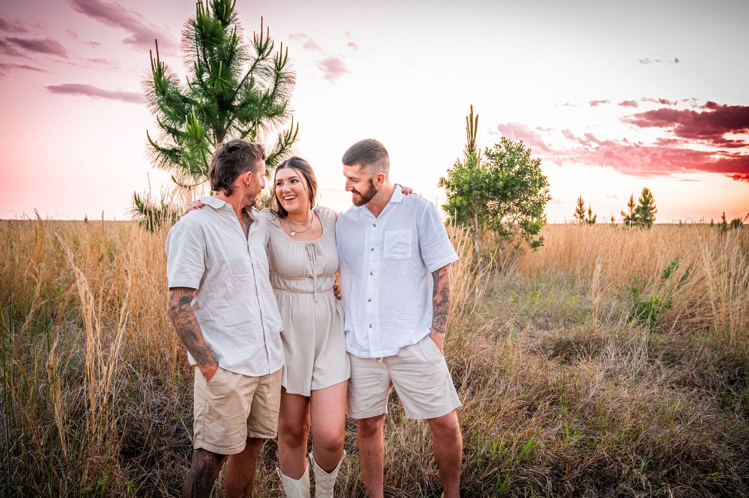 Three people dressed in light colored clothing share a laugh together in a field at sunset.