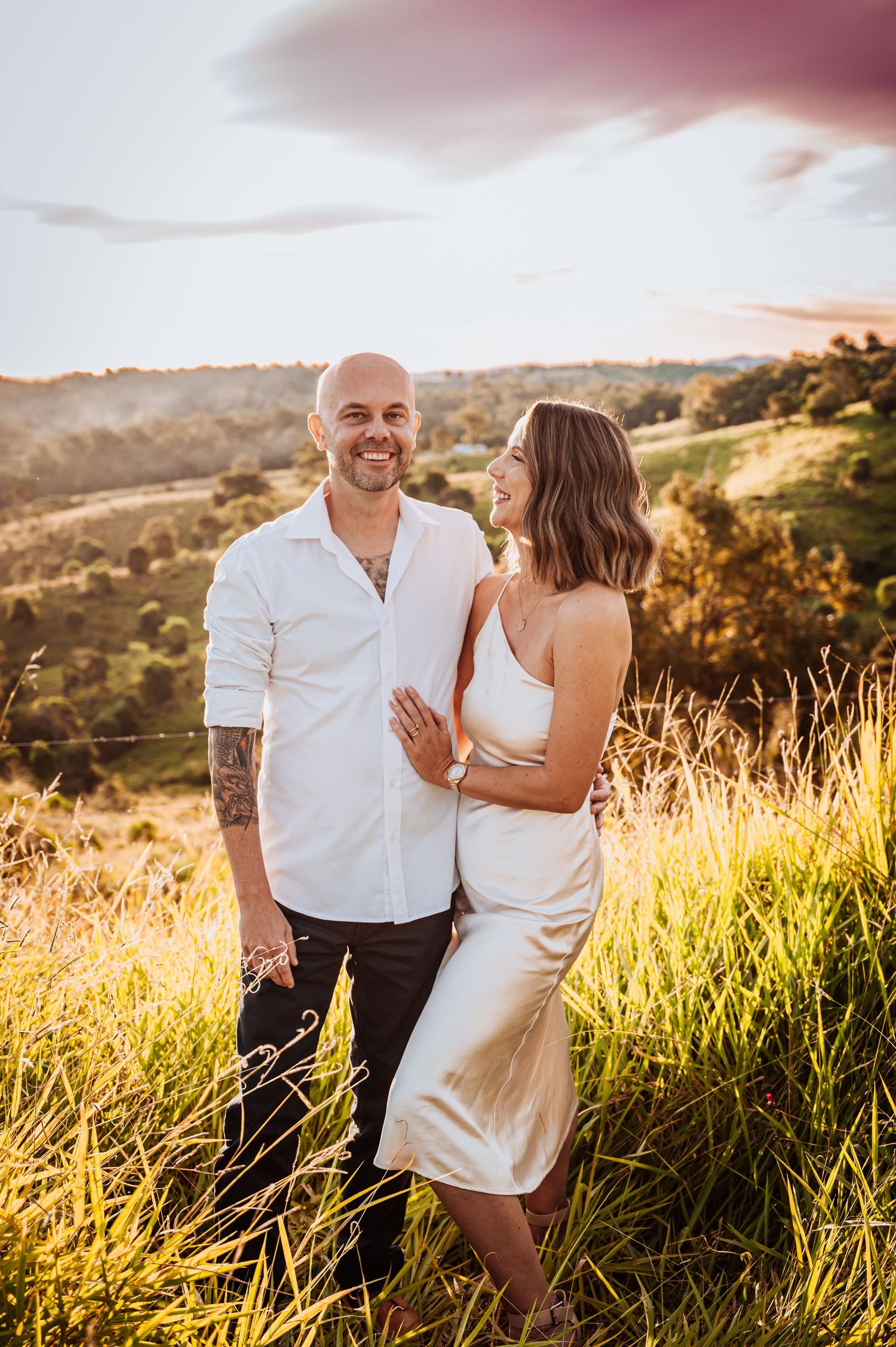 A couple laughs together while standing in golden sunlit grass on a hillside.