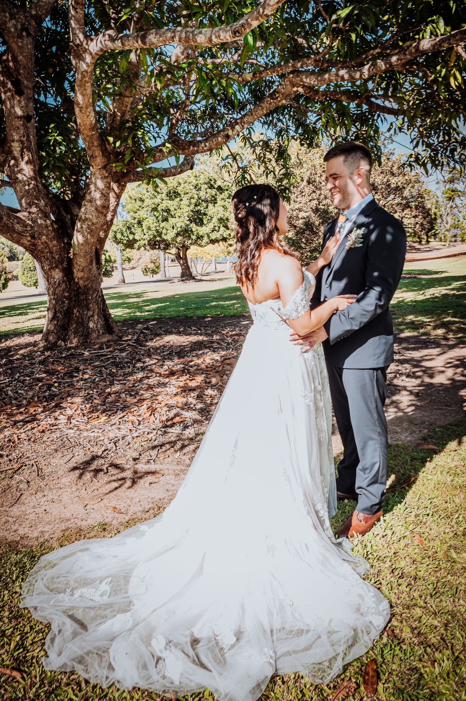A romantic wedding moment captured under a sprawling tree with a flowing white lace gown trailing behind.