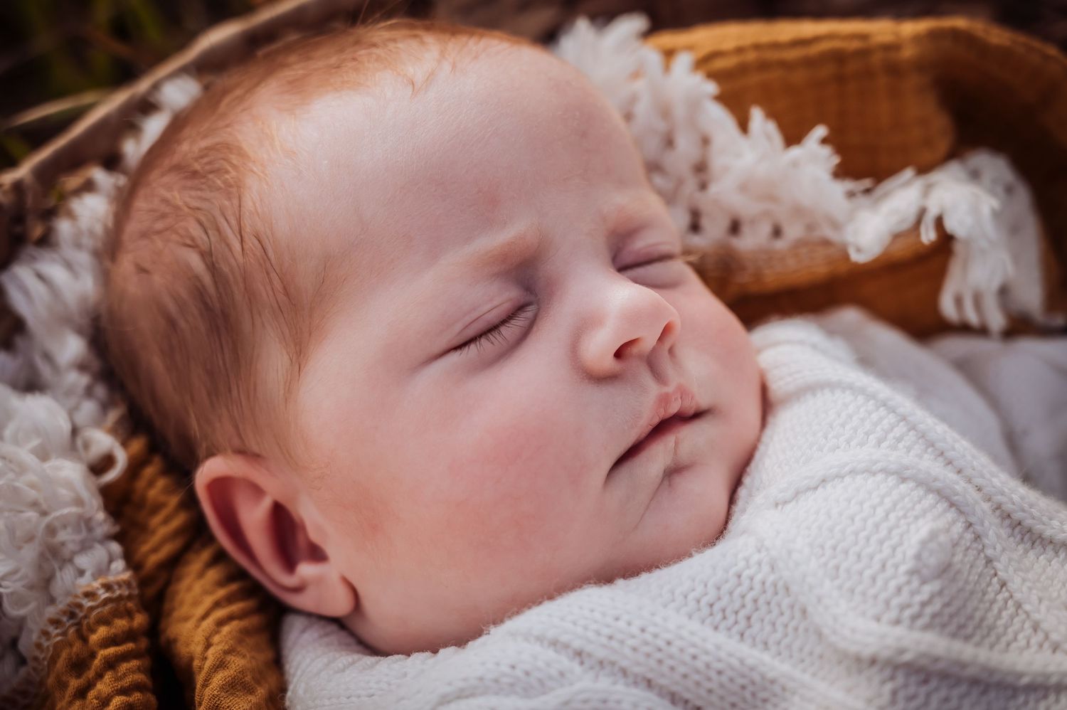 Sleeping newborn baby dressed in white knit outfit lying in woven basket.