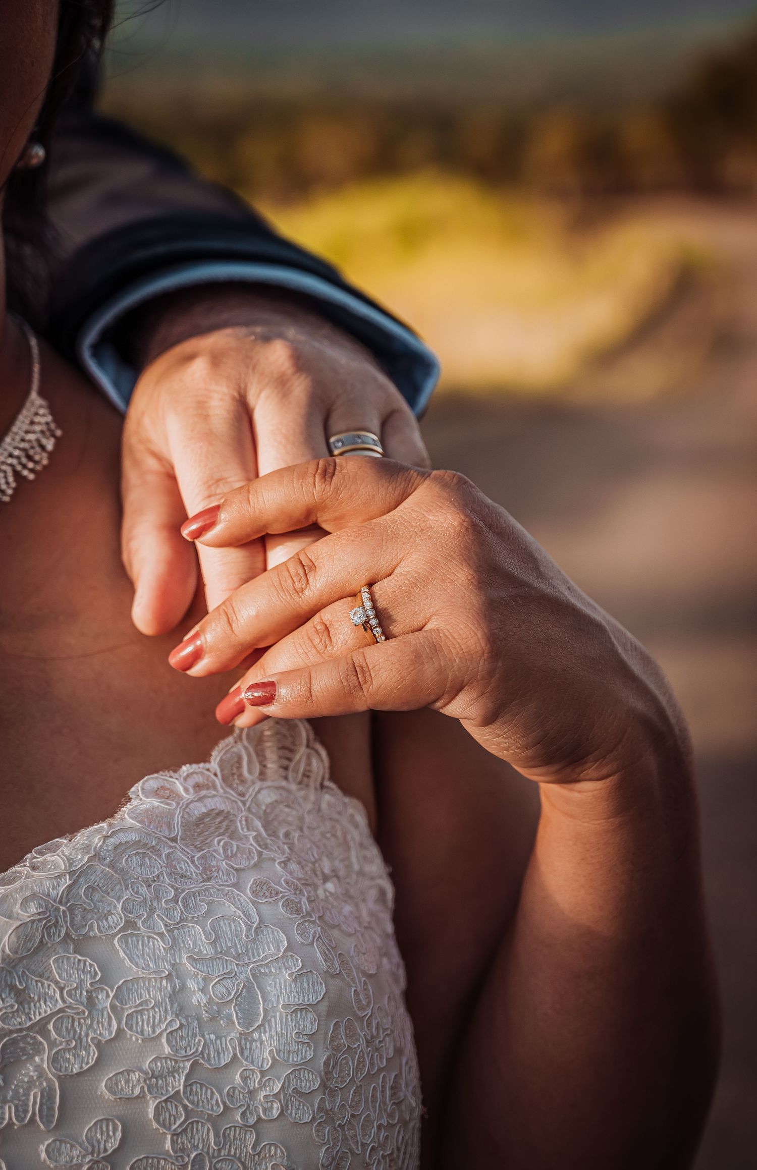 Close up of wedding rings and clasped hands showing lace dress detail in warm sunlight.