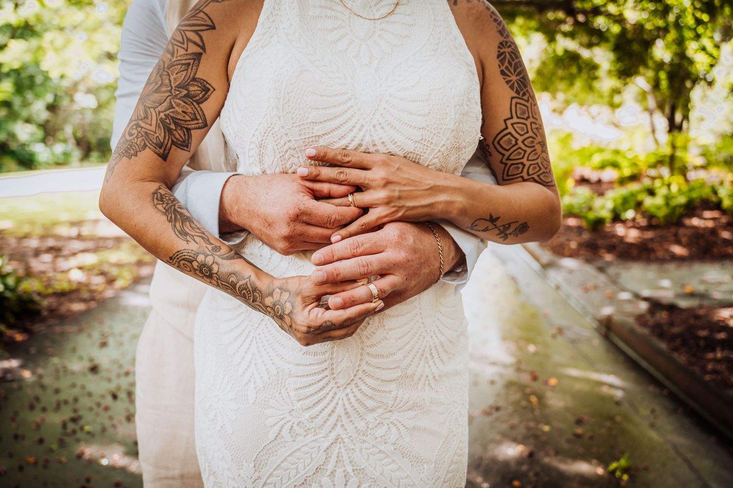 Close-up of tattooed arms and hands with wedding rings against a white lace dress.